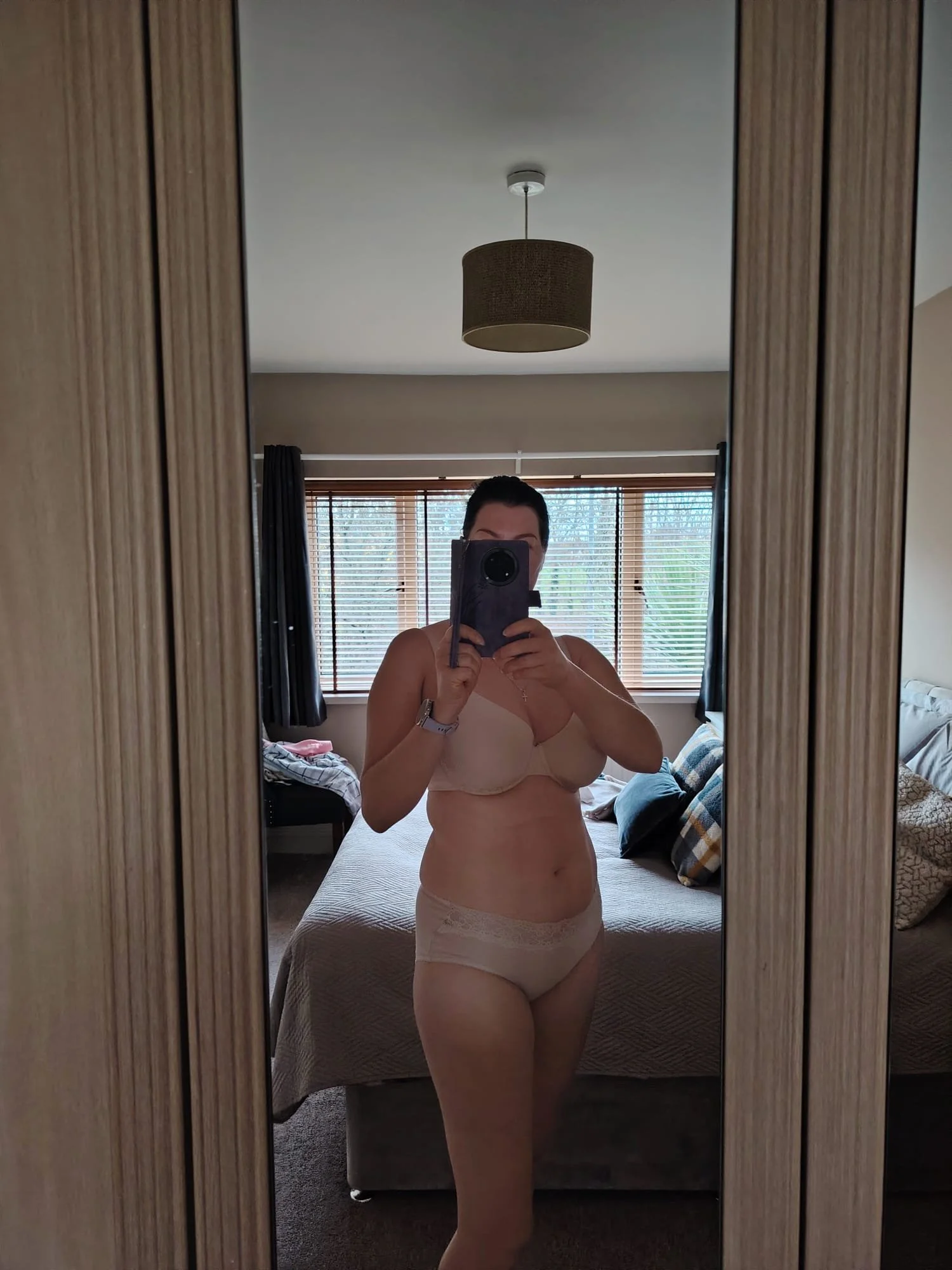 A woman taking a mirror selfie in a bedroom, wearing beige lingerie and holding a phone in front of her face. The room has a bed with pillows, a window with blinds, and a hanging ceiling light.
