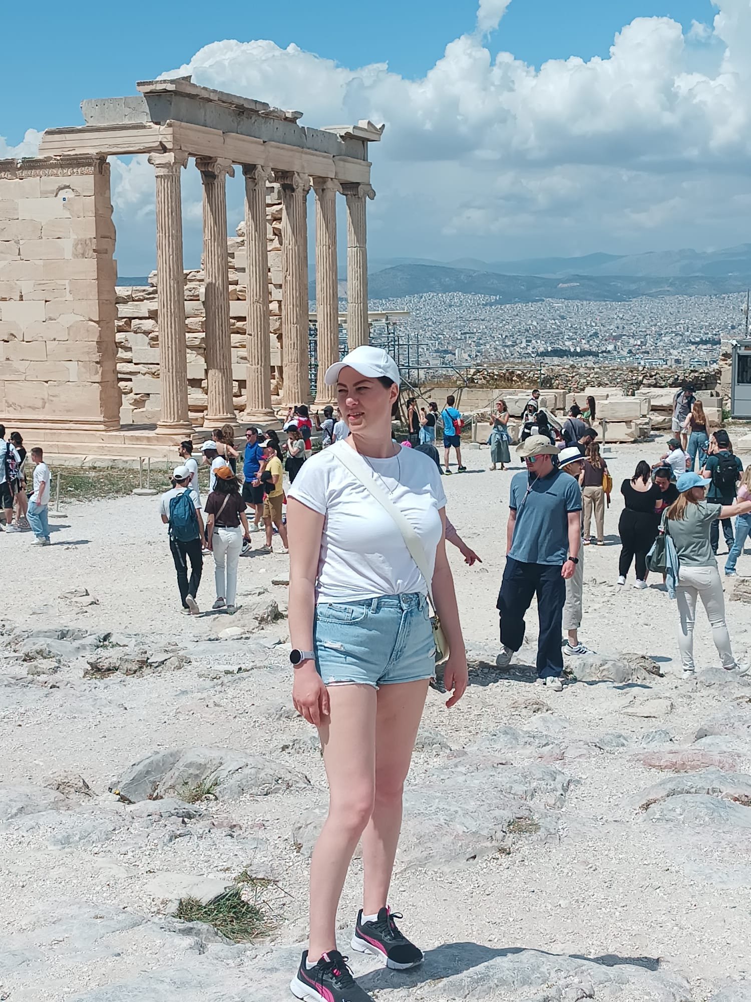A woman wearing a white cap, white t-shirt, light blue shorts, and black sneakers standing on a rocky ground at the Acropolis in Athens, Greece. Ancient ruins and the cityscape of Athens with mountains are visible in the background on a sunny day with clouds.