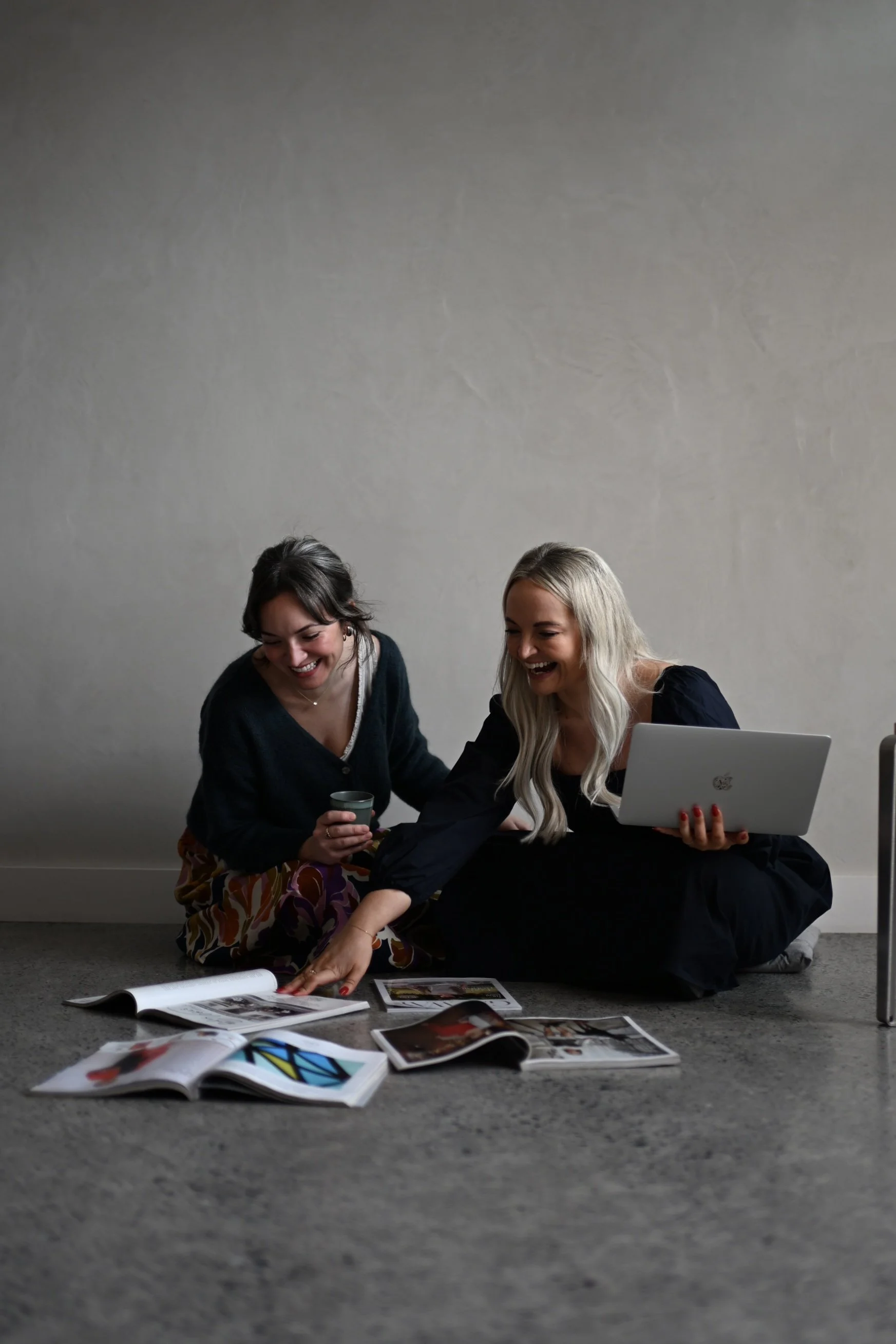 Two sisters sitting on the floor surrounded by social strategy, one holding a coffee mug and the other using a laptop, both looking at creative content and smiling.