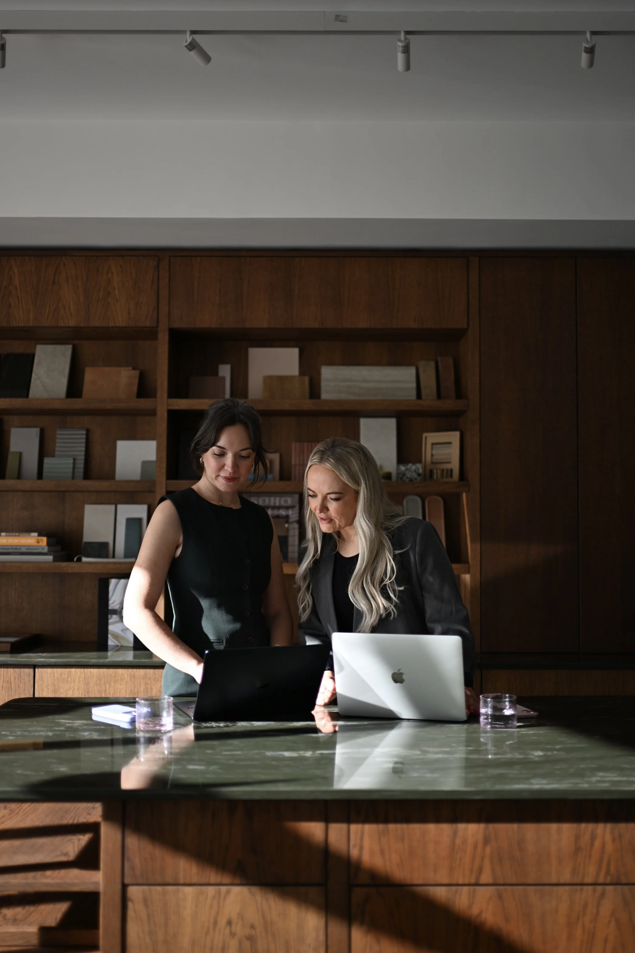 Two women in black outfits looking at a laptop and a tablet in a modern office or conference room with wooden shelves and a green marble table.