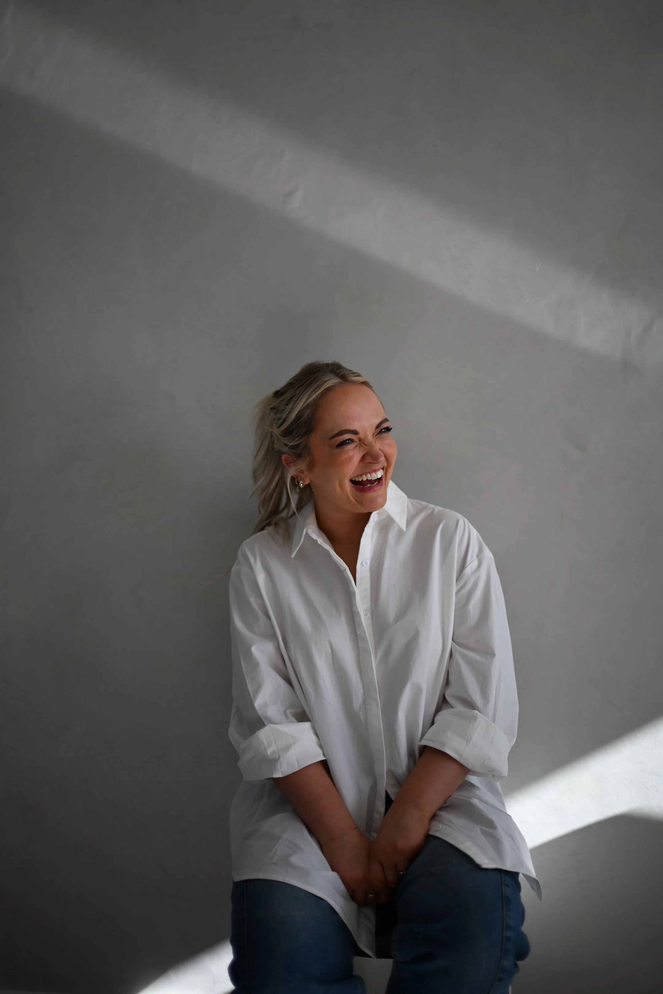 A social strategist with blonde hair in loose waves, wearing a white button-up shirt and blue jeans, is sitting on a stool against a plain, light grey wall. She is laughing or smiling broadly with her eyes closed and her head slightly tilted back.