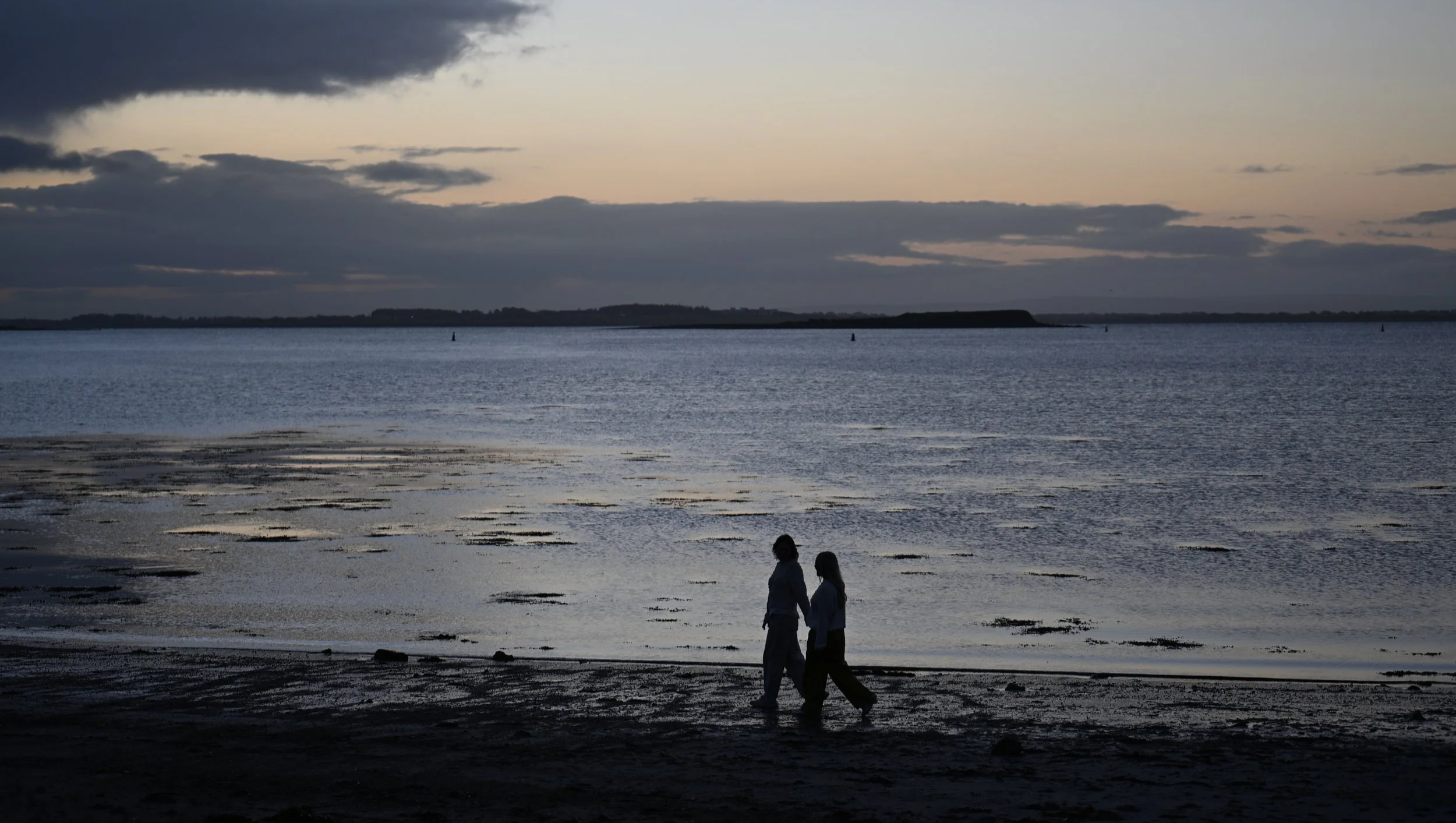 Two people holding hands and walking along a beach at sunset with calm water and clouds in the sky.