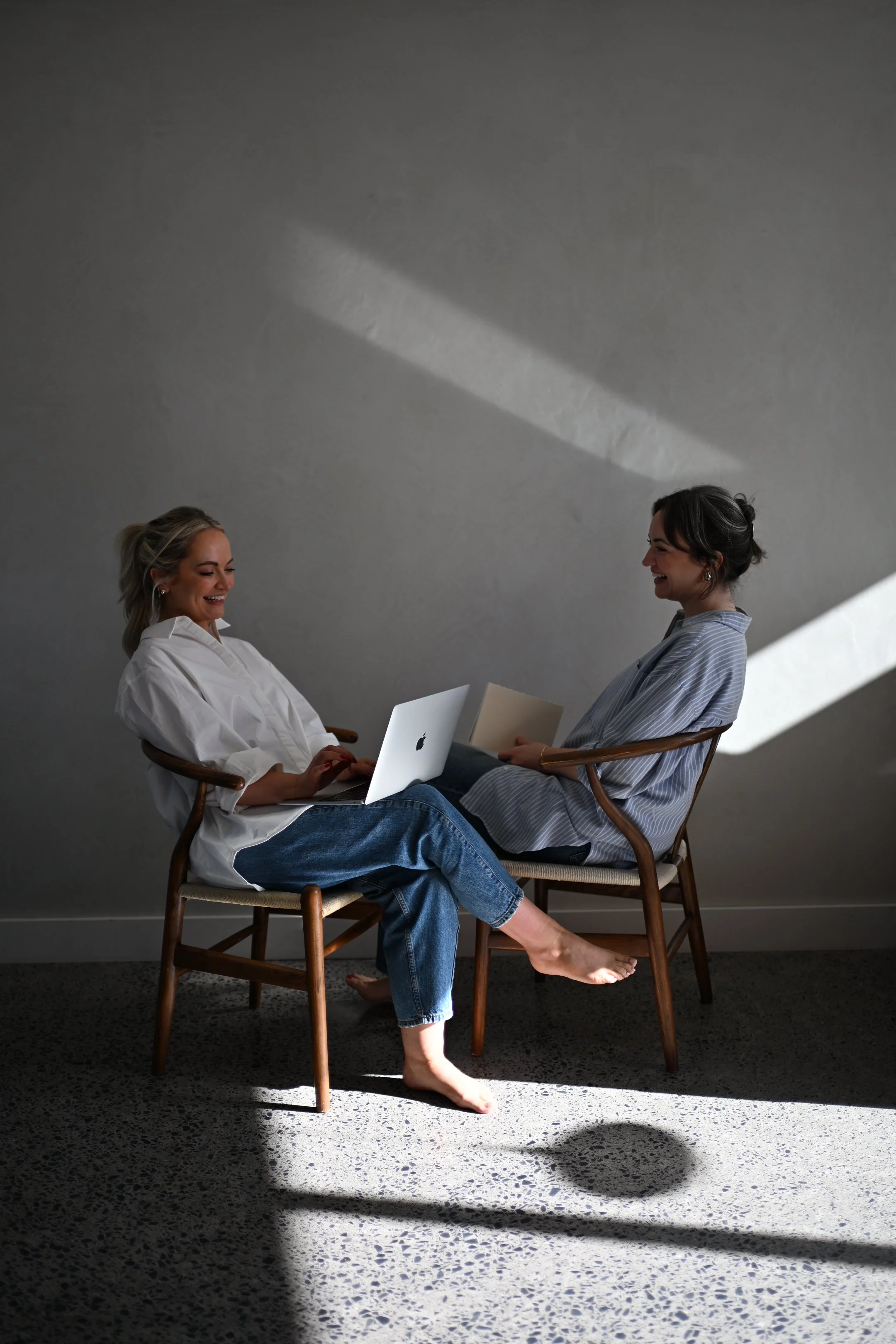 Two social media experts in casual clothing sitting on wooden chairs facing each other, smiling and engaging in a conversation. One woman working on creative and the other on research.