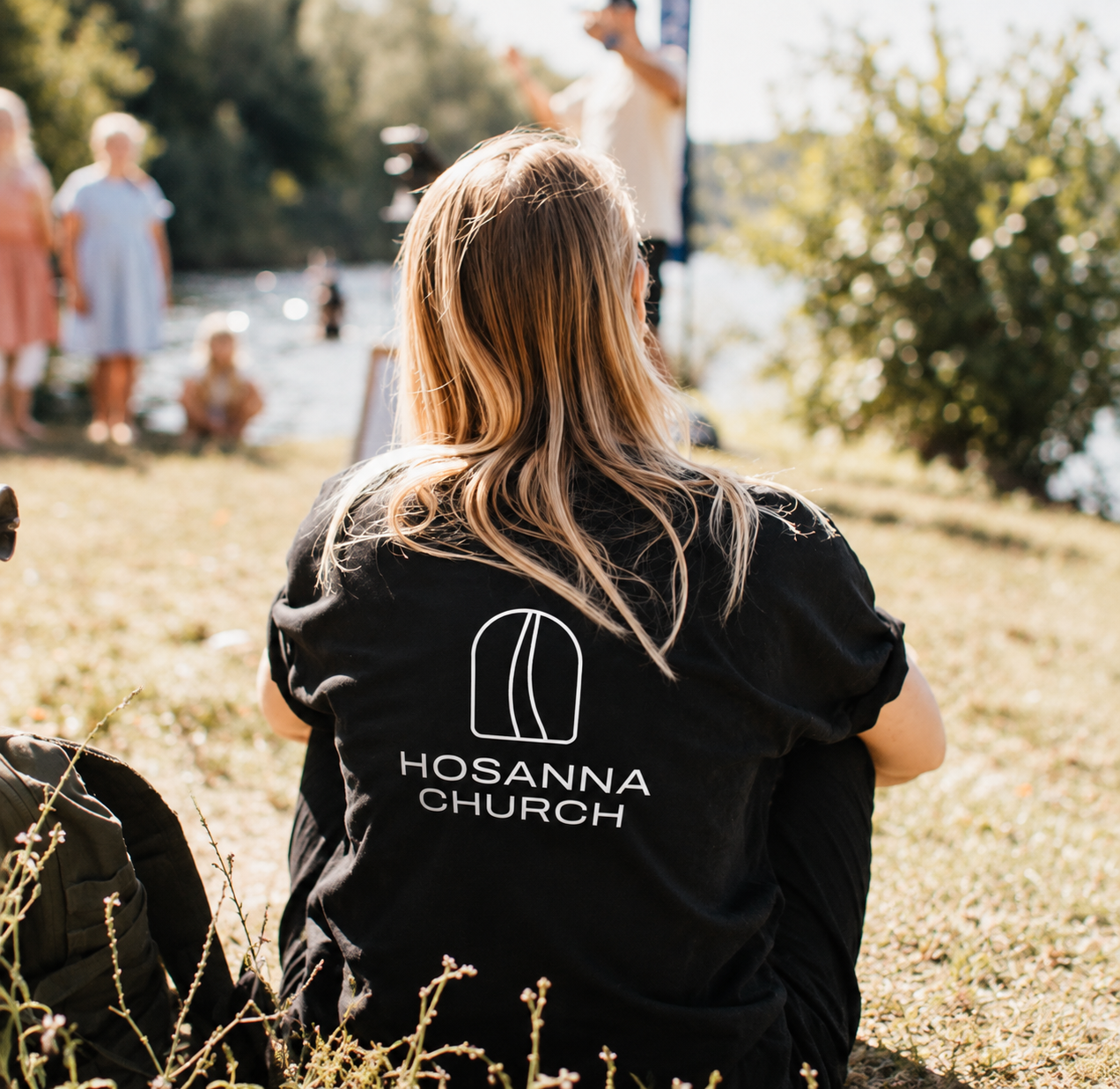 Frau sitzt im Freien mit einem schwarzen T-Shirt mit dem Logo 'Hosanna Church' auf dem Rücken, während im Hintergrund Menschen an einem Wasserufer sind.