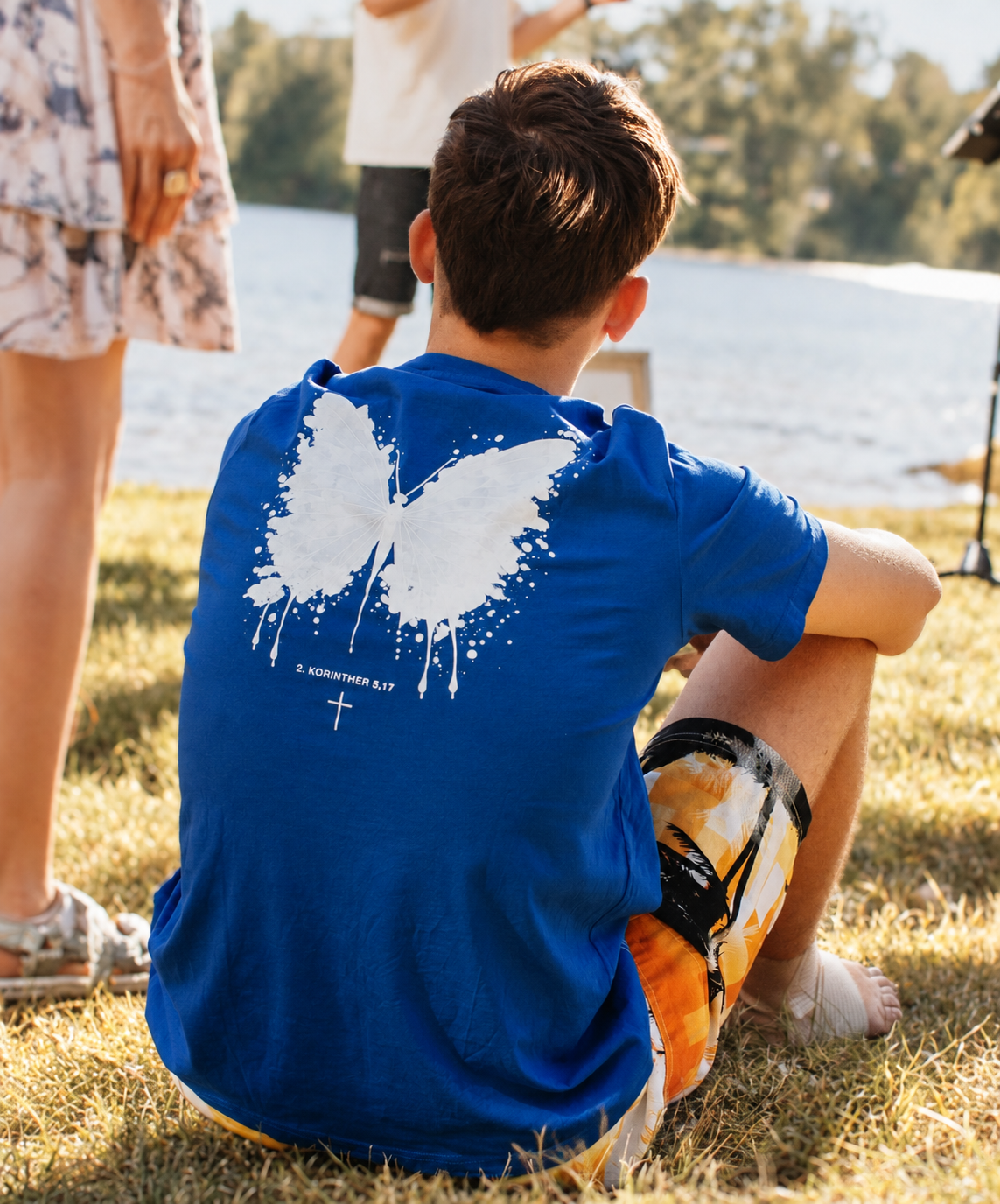 Junge sitzt am Flussufer, trägt ein blaues T-Shirt mit einem weißen Schmetterlingsdesign und bibelverse, umgeben von anderen Personen bei sonnigem Wetter.