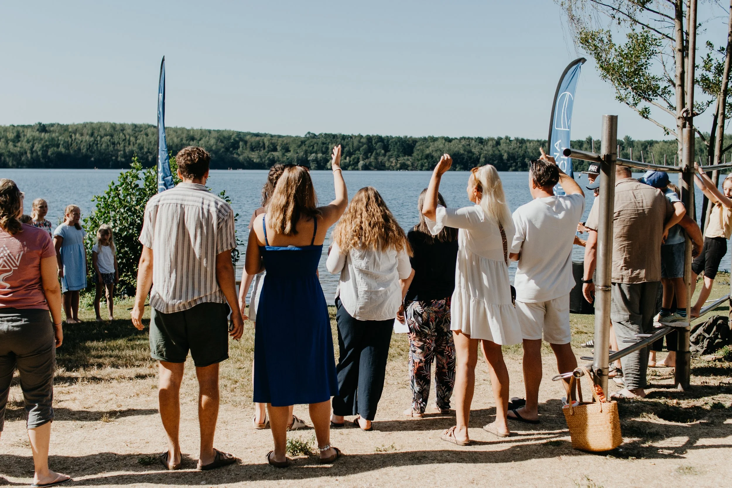 Menschen am See, einige heben die Hände, sitzen, sprechen, im Freien an einem sonnigen Tag.