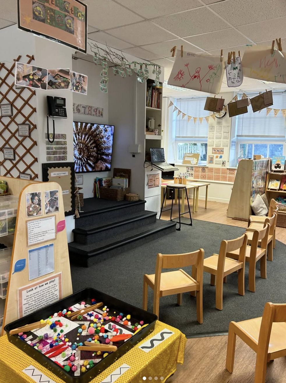 A classroom with small wooden chairs arranged in a semi-circle, a table with art supplies, storage units, and colorful children's artwork hanging from a clothesline. There are windows, a TV screen, and educational posters.