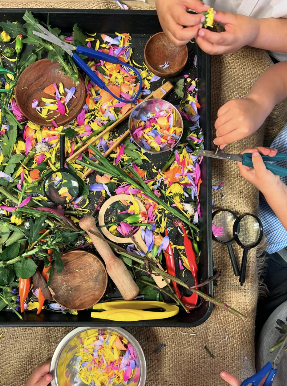 Children's hands using scissors to trim colorful flower petals during a craft activity; workspace is filled with vibrant petals, green leaves, and various crafting tools.