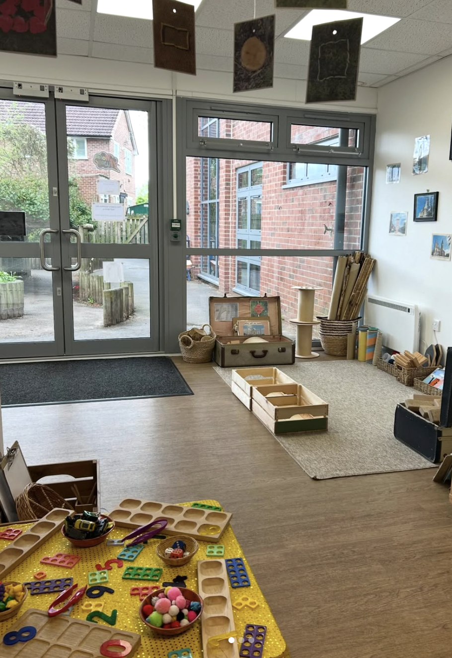 Inside a classroom with educational toys, wooden learning materials, and artwork on the walls. Large glass doors and windows reveal an outdoor area with plants, trees, and brick buildings.