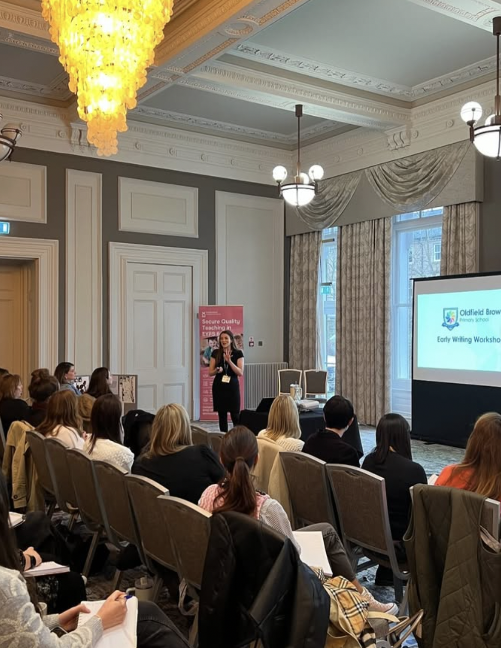 A woman speaking at a conference in a decorative room with chandeliers, large windows with curtains, and a screen displaying a presentation titled 'Early Welling Workshop' at Oldfield Brow.