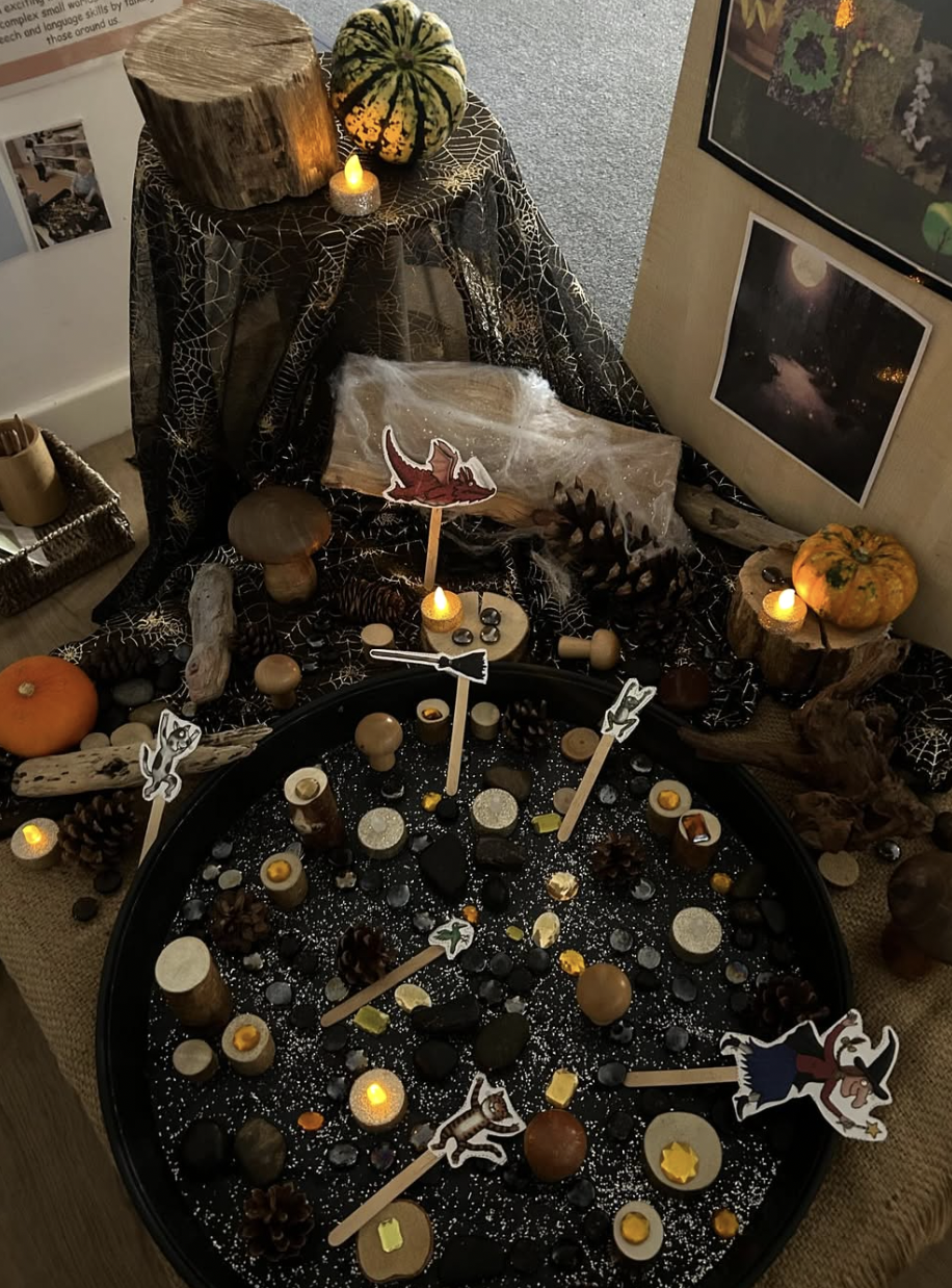 A Halloween-themed display with a black tray filled with stones, miniature pumpkins, pinecones, and decorations, illuminated by small candles. Surrounding the tray are pumpkins, wooden tree slices, a spider web cloth, and Halloween-themed cutouts of witches and bats. There's a wall with pictures and a framed scene of a moonlit landscape.
