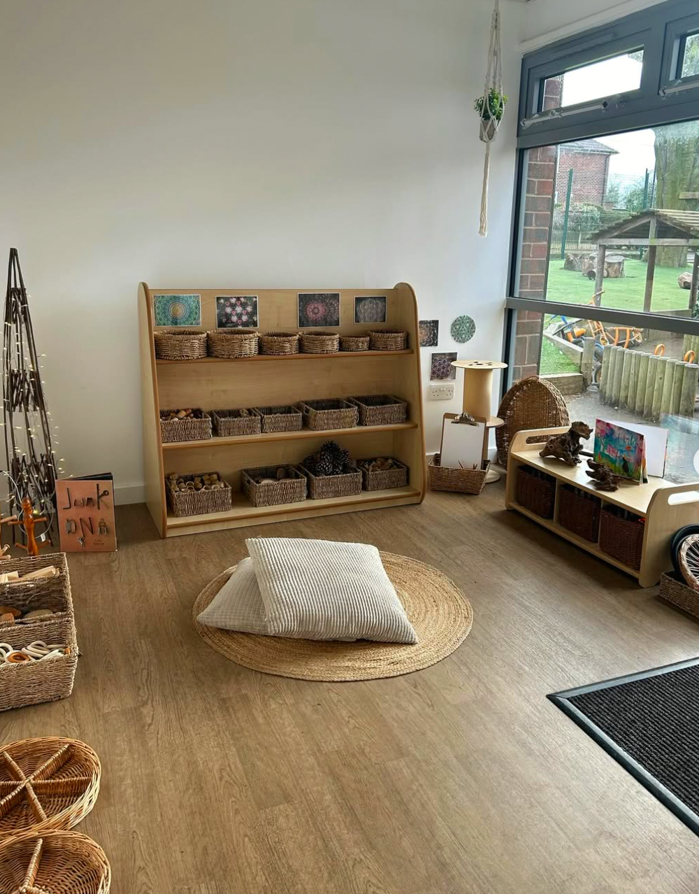 A cozy playroom with a wooden shelf filled with wicker baskets, a round woven rug with pillows, and outdoor scenery visible through large windows.