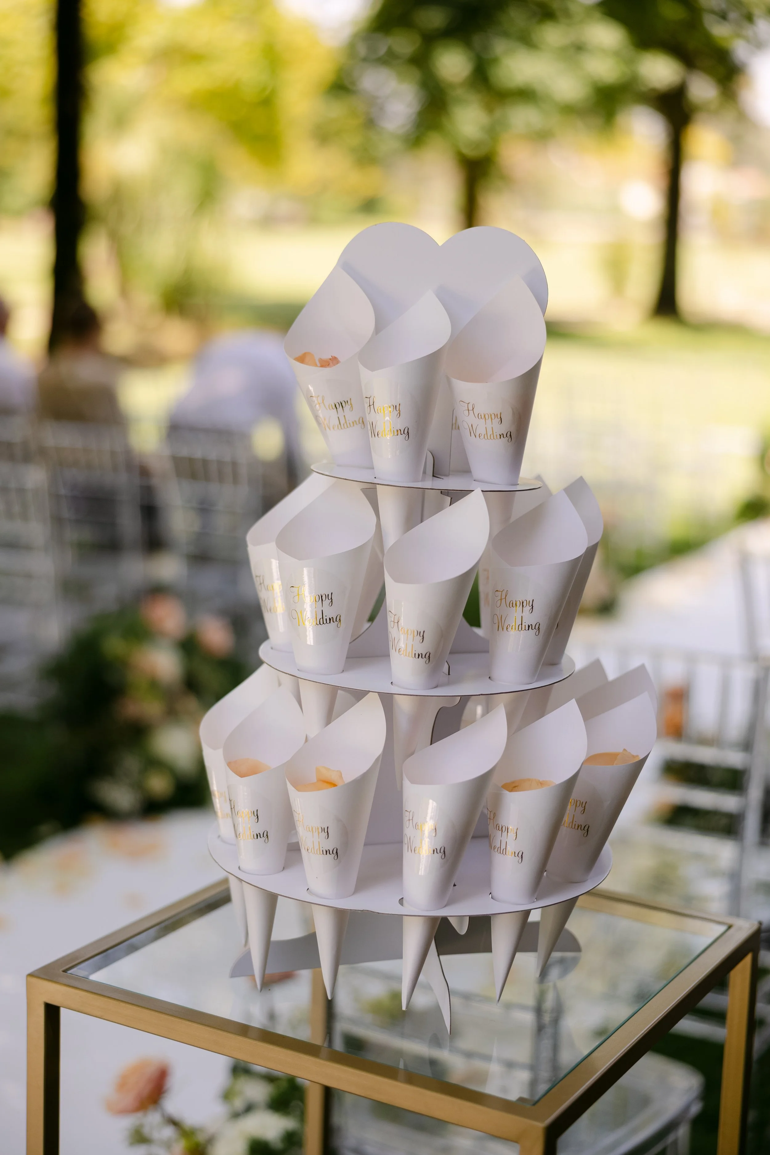 Torre de convites de casamento em formato de cone branco com mensagem "Happy Wedding" em dourado, decorando uma mesa em acara ao ar livre com árvores ao fundo.