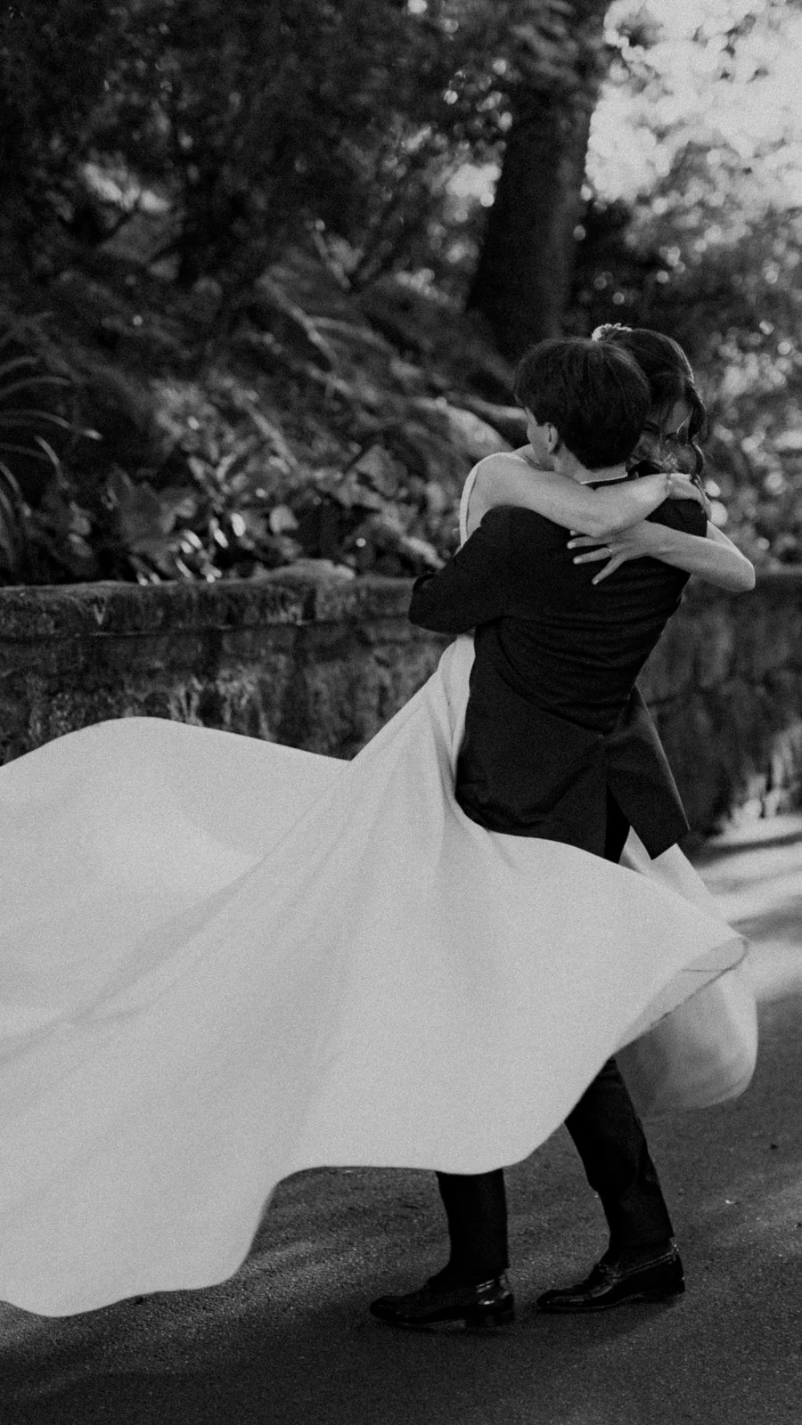 Casal se abraçando, fotografados em preto e branco ao ar livre, possivelmente em um momento romântico ou de casamento.