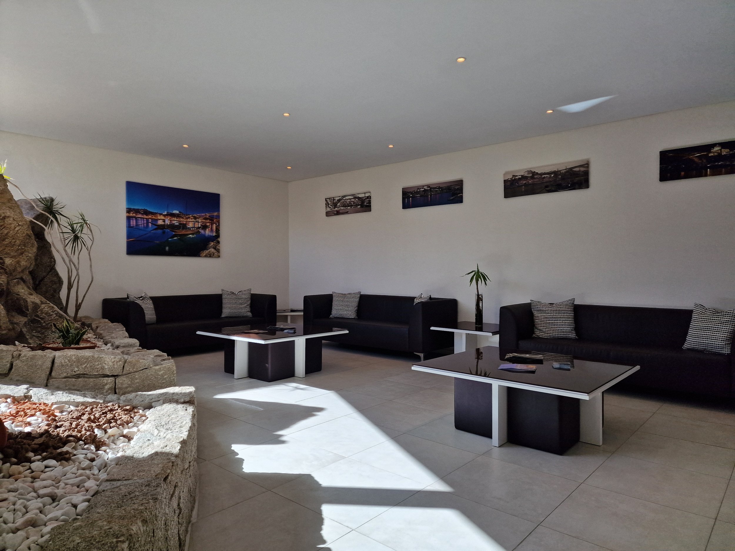 Sala de estar moderna com sofá preto, quadros de paisagens urbanas nas paredes, plantas e uma área decorativa com pedras e plantas na esquerda.