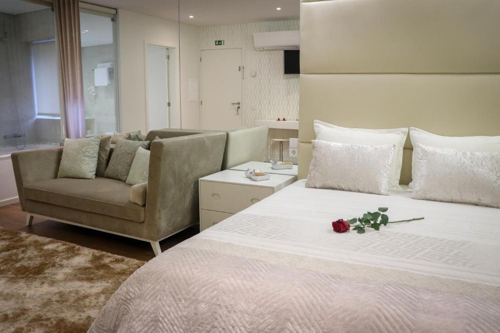 Quarto de hotel com cama de casal, travesseiros brancos e uma rosa vermelha sobre ela, sofá cinza com almofadas, mesas de cabeceira brancas e decoração moderna.