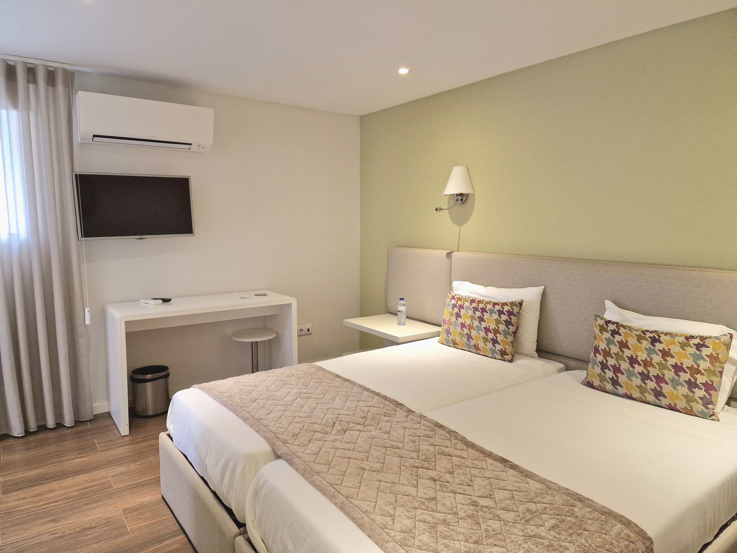Quarto de hotel com duas camas de casal com roupas de cama brancas, travesseiros com estampas coloridas, parede de cor verde claro, televisão fixa na parede à esquerda, ar-condicionado acima da TV, luminária de parede ao lado da cama, mesa ao lado da