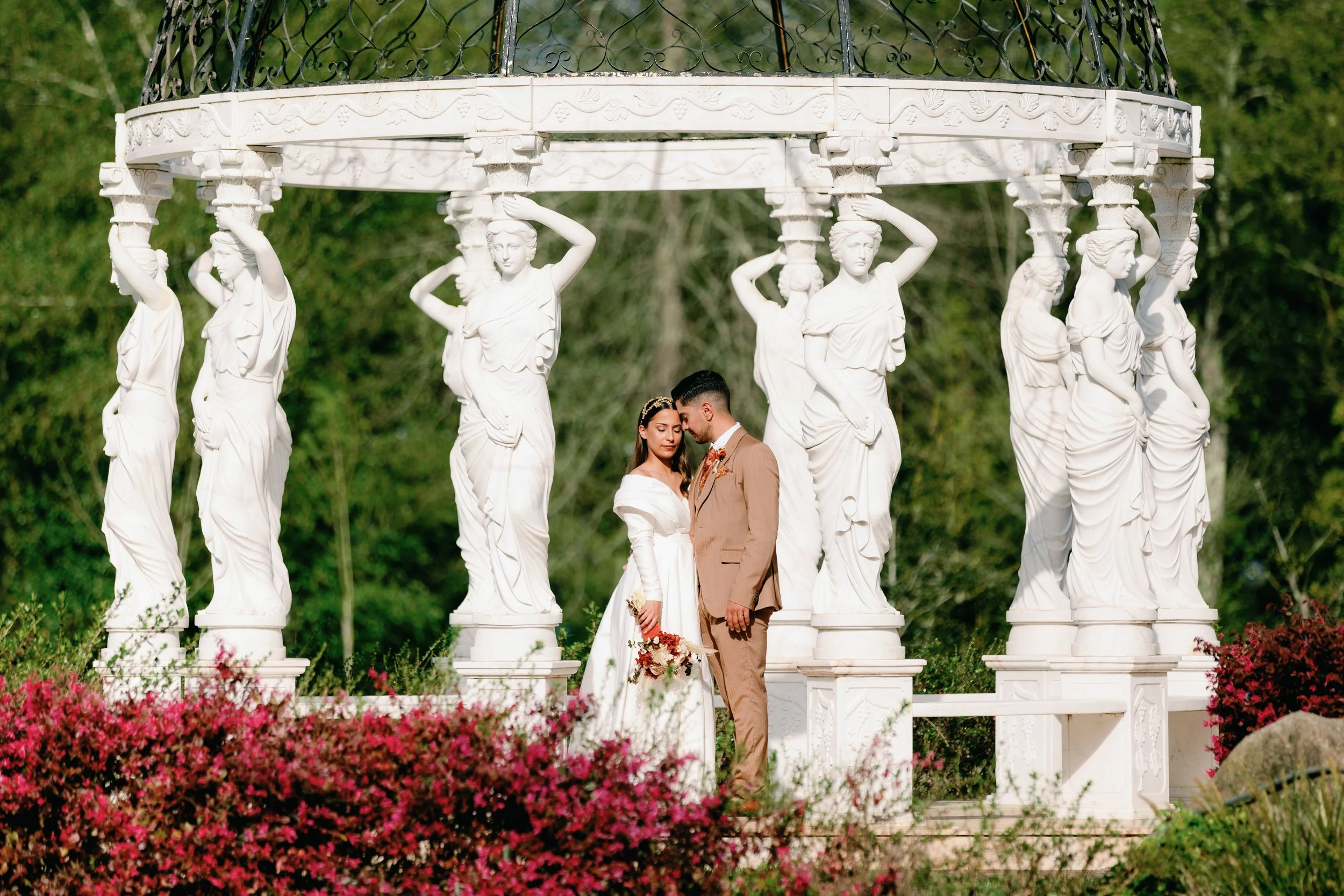 Casal de casamento com vestido branco e terno bege, deitados diante de uma estrutura decorativa com estátuas de mulheres neoclássicas segurando a moldura superior, cercados por flores coloridas e vegetação ao fundo.