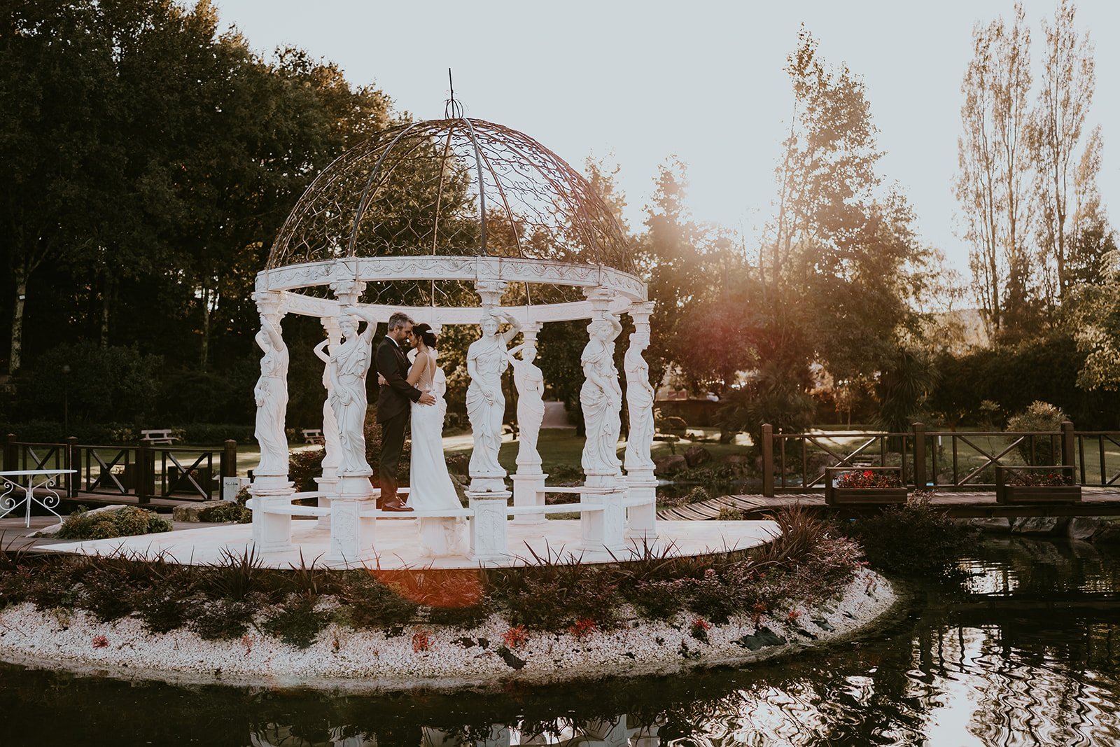 Casal de casamento em uma cerimônia ao ar livre na igreja clássica branca com colunas e cúpula decorativa, ao entardecer, ao lado de um lago cercado de árvores.