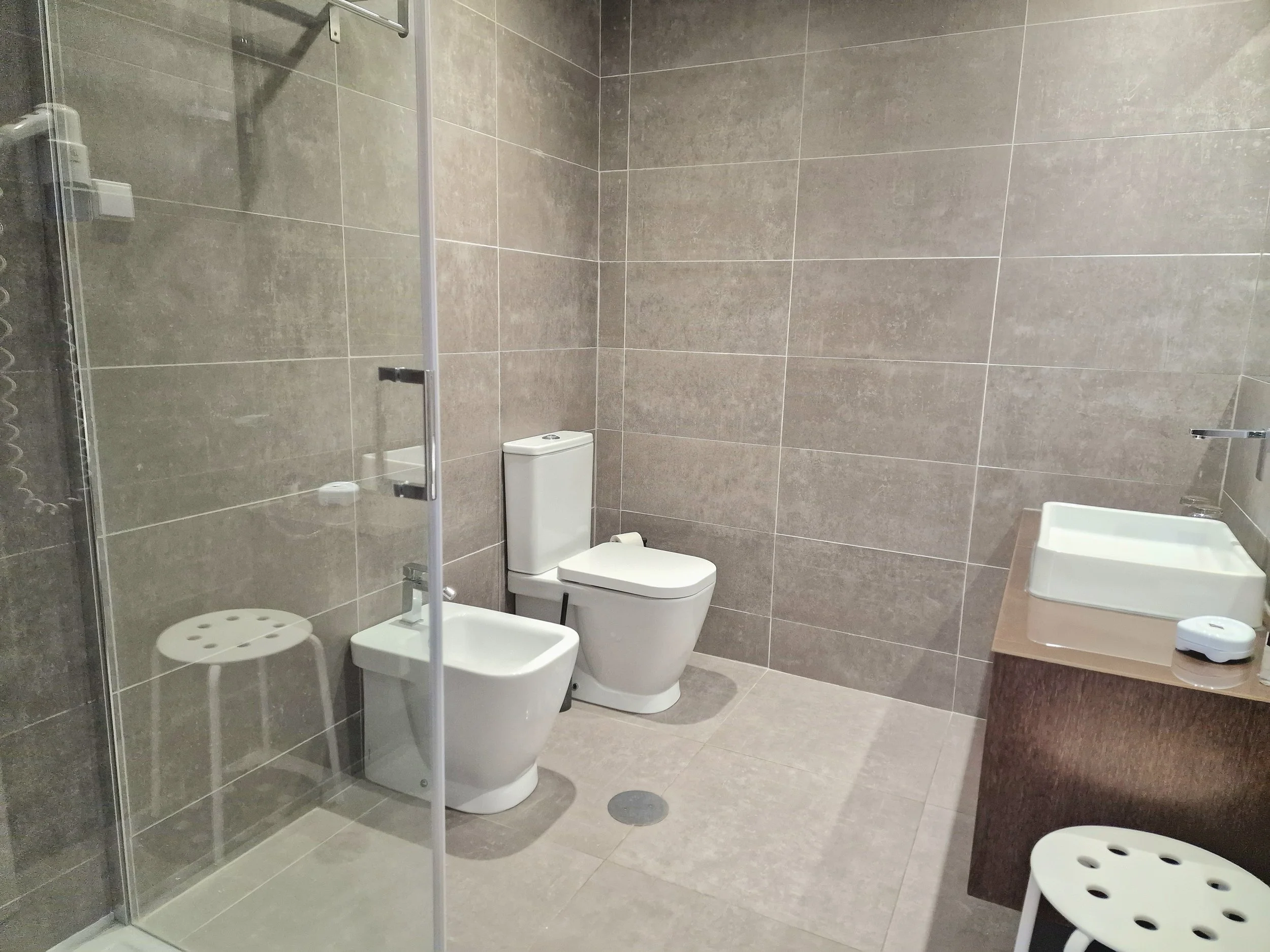 Banheiro com paredes e chão de revestimento cinza, com vaso sanitário branco, pia com bancada de madeira e espelho acima, ducha e cadeira de plástico branco na área do chuveiro.