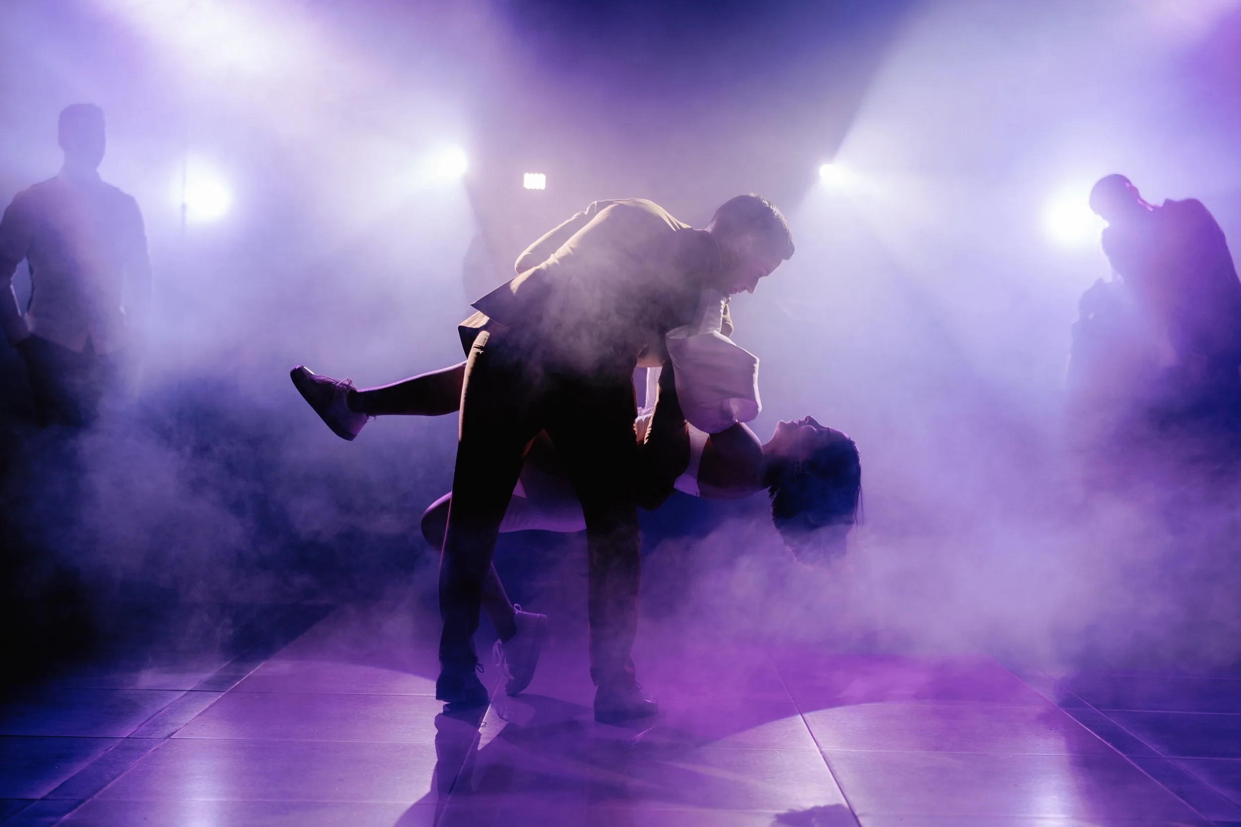 Casal dançando no palco sob luzes coloridas com fumaça e sombras ao fundo.