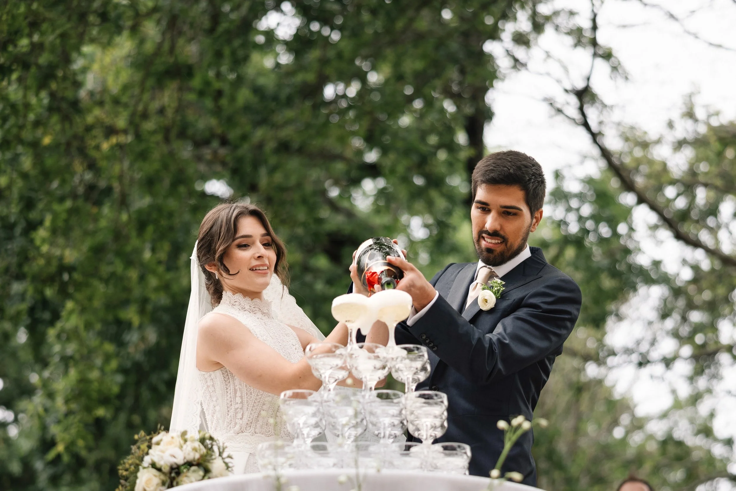 Casal de noivos servindo champanhe em um jardim durante festa de casamento ao ar livre.