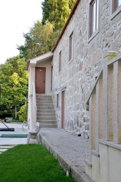 Casa de pedra com escada de concreto, janelas pequenas e uma porta em um dos andares, em uma área com árvores verdes.