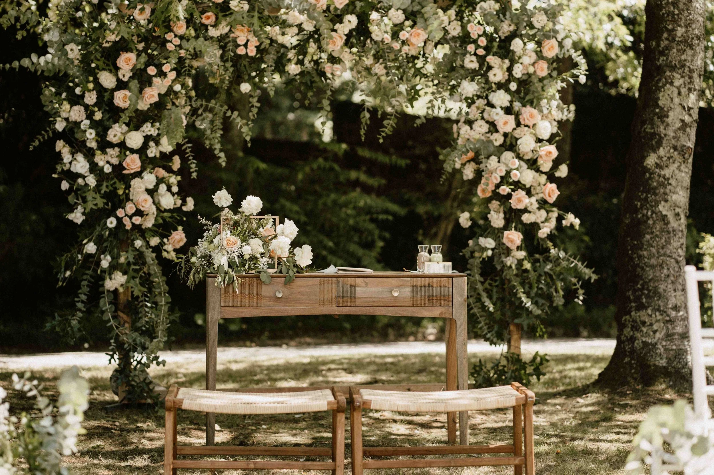 Decoração de casamento ao ar livre com arco feito de flores brancas e rosas pêssego, decorando uma mesa de madeira com duas cadeiras de palha ao nível do solo em um ambiente cercado de árvores.