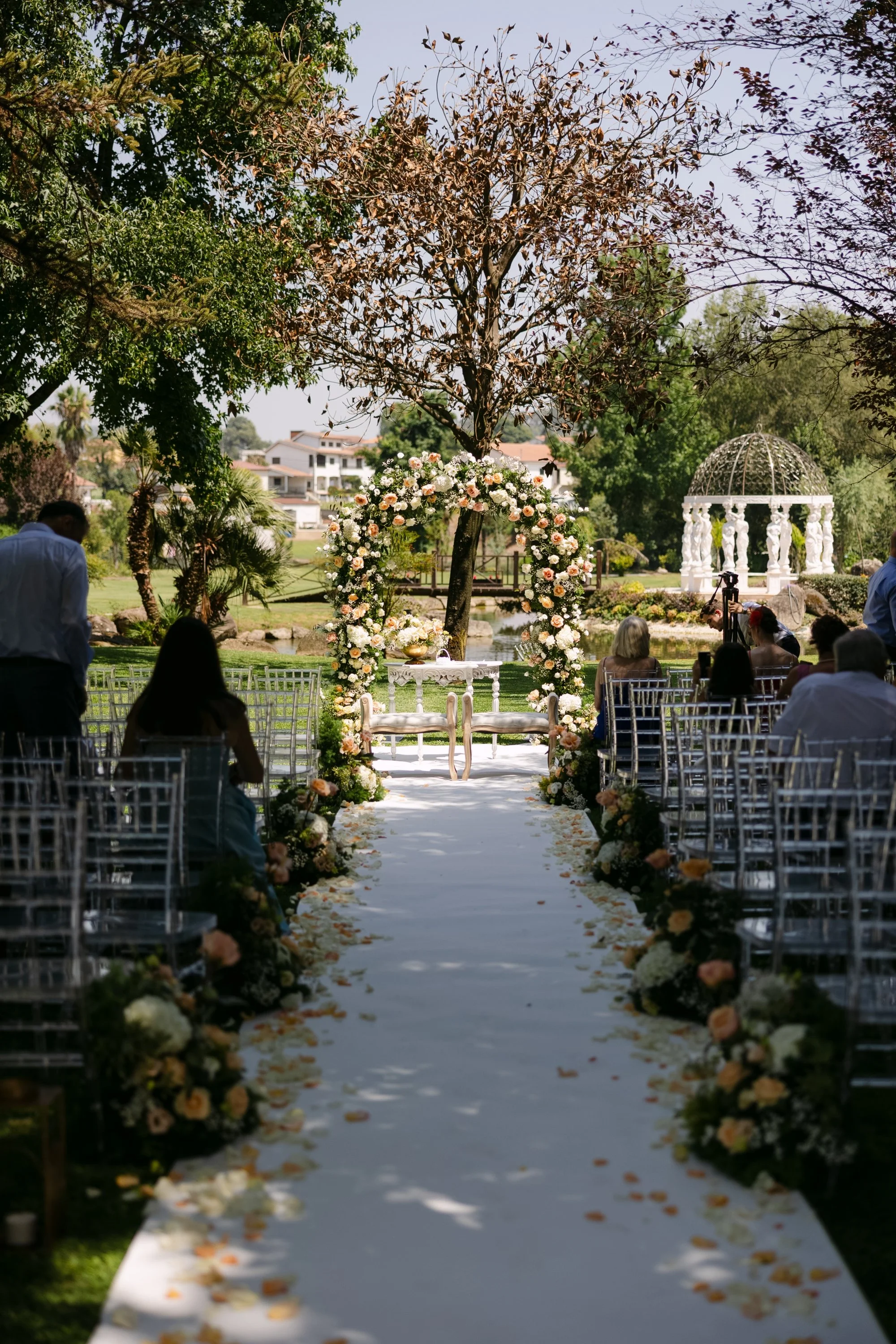 Cerimônia de casamento ao ar livre, com decoração de flores em arco e em volta do caminho, árvores ao redor, e uma estrutura branca ao fundo com pessoas sentadas assistindo.