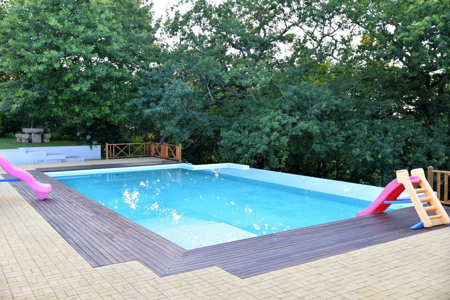 Piscina em um arredores com área de deck de madeira, cercada por árvores verdes, com escorregadores infantis rosas ao lado da piscina.