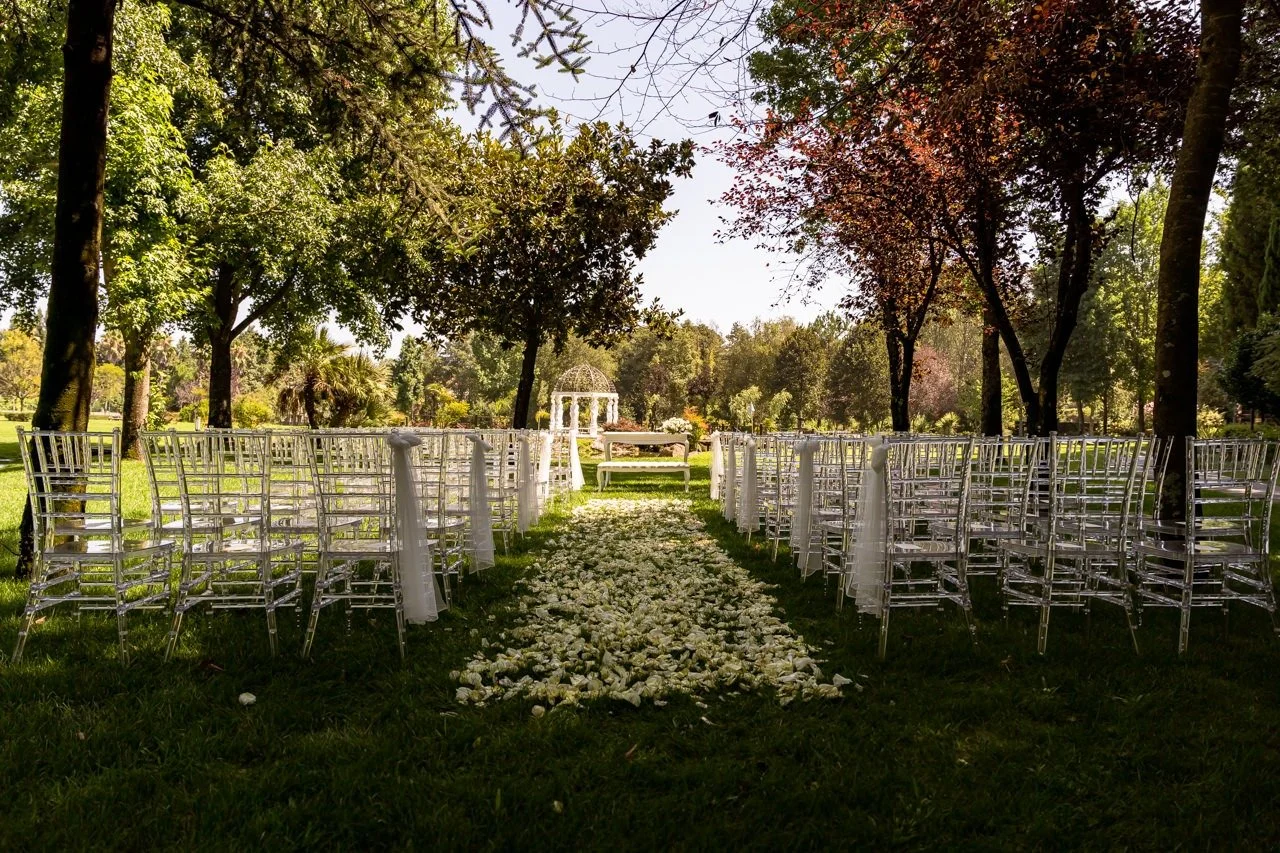 Cerimônia ao ar livre em um jardim com cadeiras brancas e uma passarela de pétalas de rosa, com uma estrutura de gazebo no fundo cercada por árvores verdes.