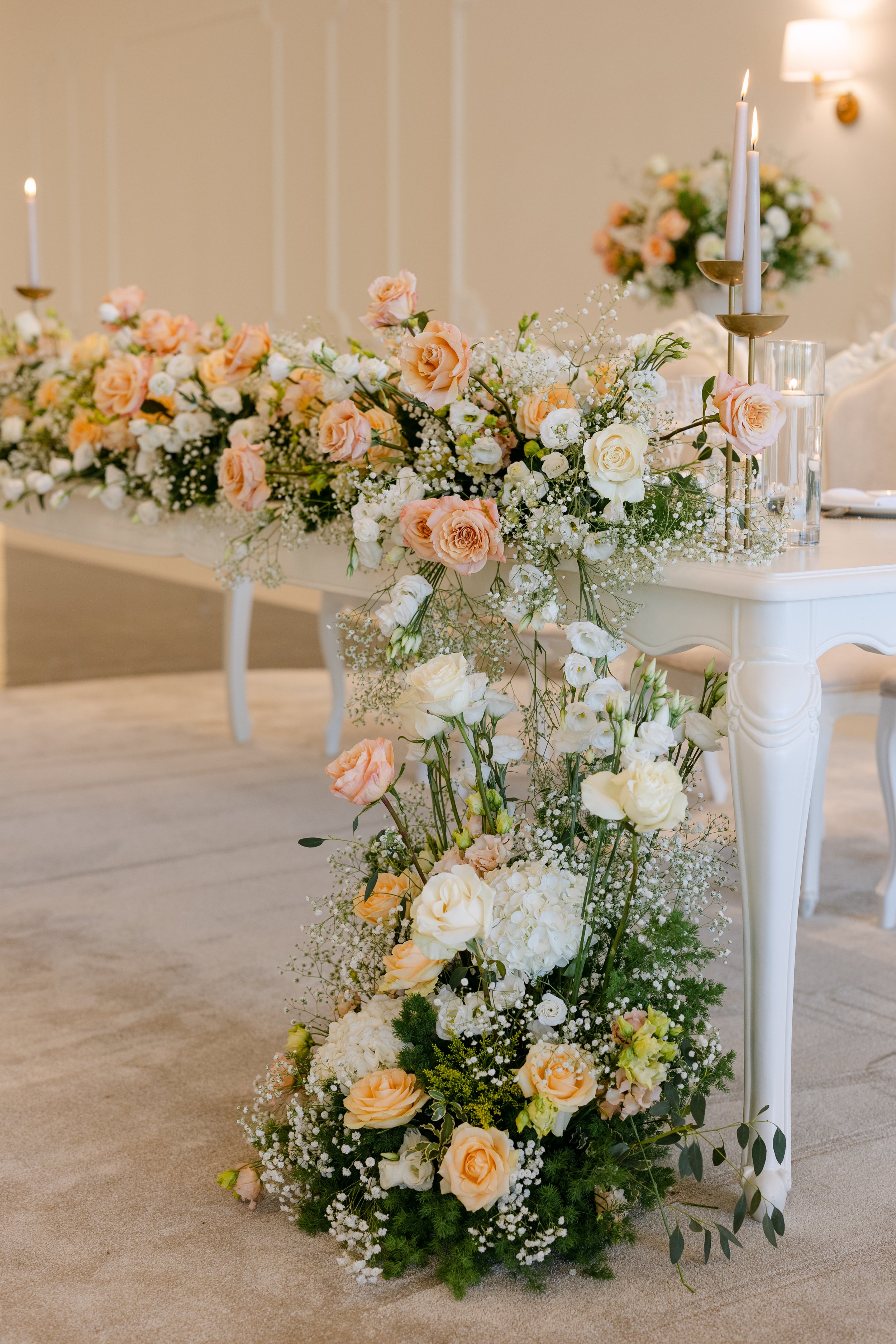 Arranjo florido de casamento com rosas brancas e rosas cor de pêssego, acompanhado de galhos verdes e pequenos buquês brancos, decorando uma mesa elegante com velas e castiçais dourados, em ambiente com parede clara e iluminação suave.