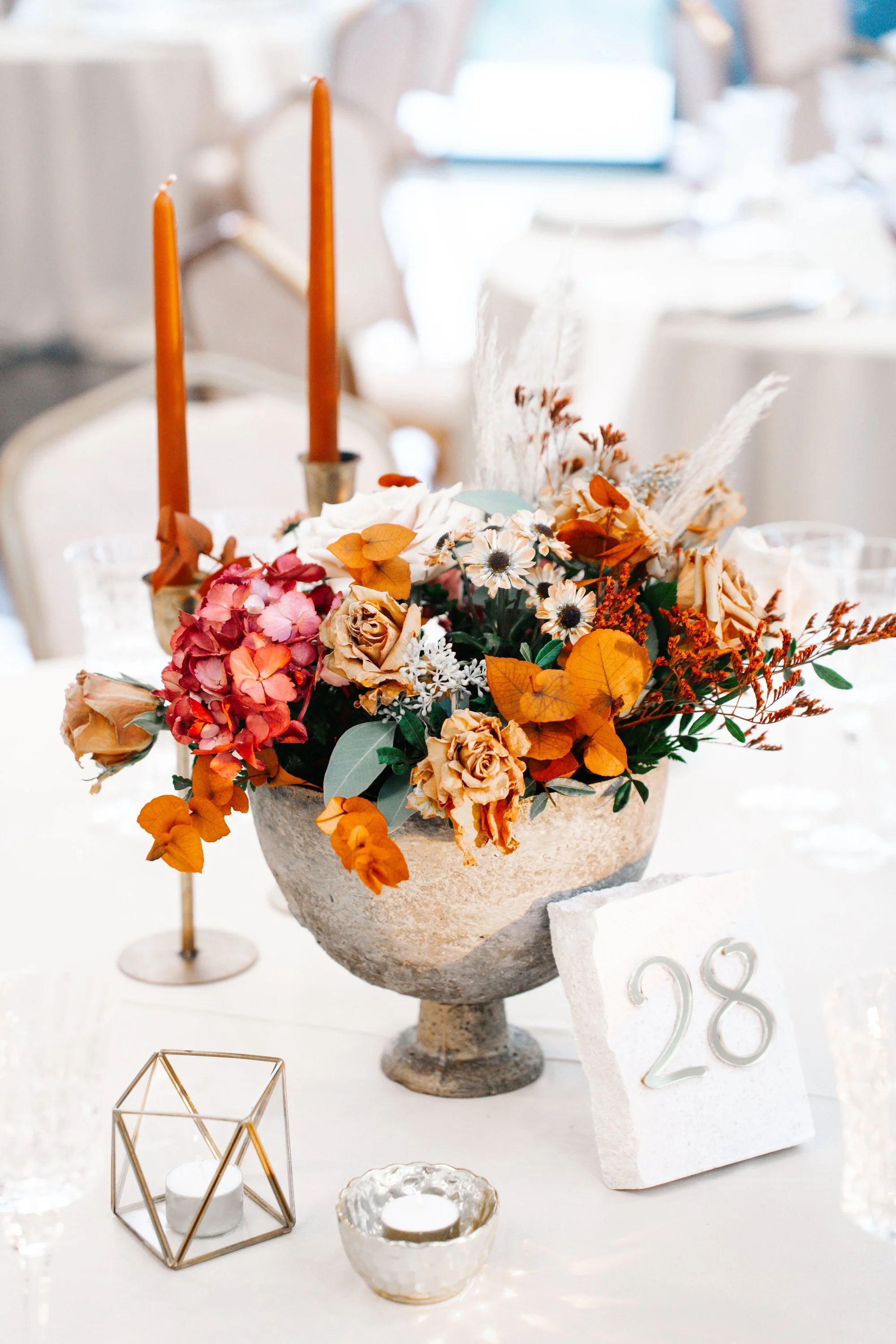 Arranjo de flores com rosas, gérberas e folhas em vaso de pedra decorado com velas e enfeites de decoração dourados e brancos, indicado para evento ou festa.