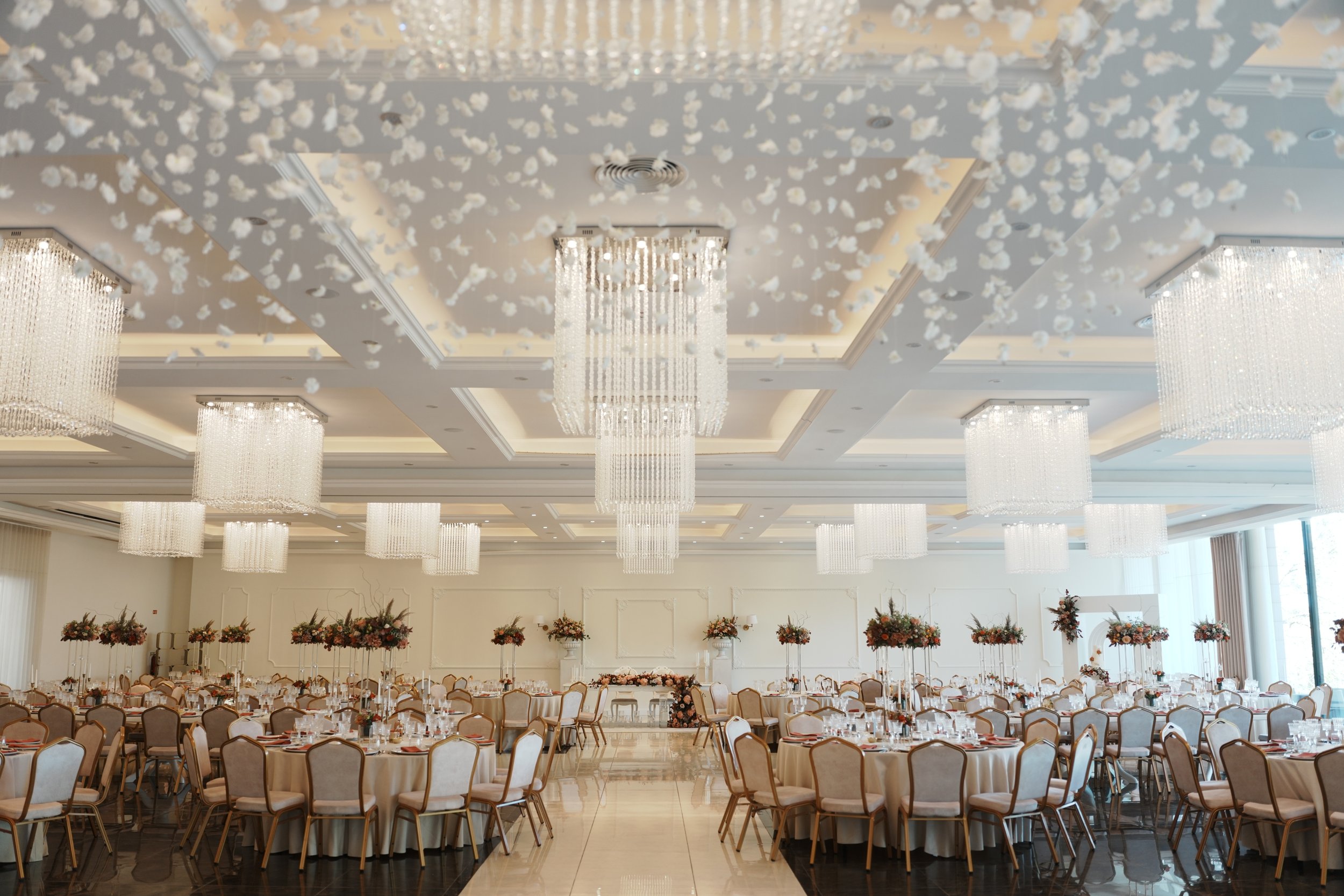 Salão de festa decorado com lustres de cristal e mesas redondas com flores.