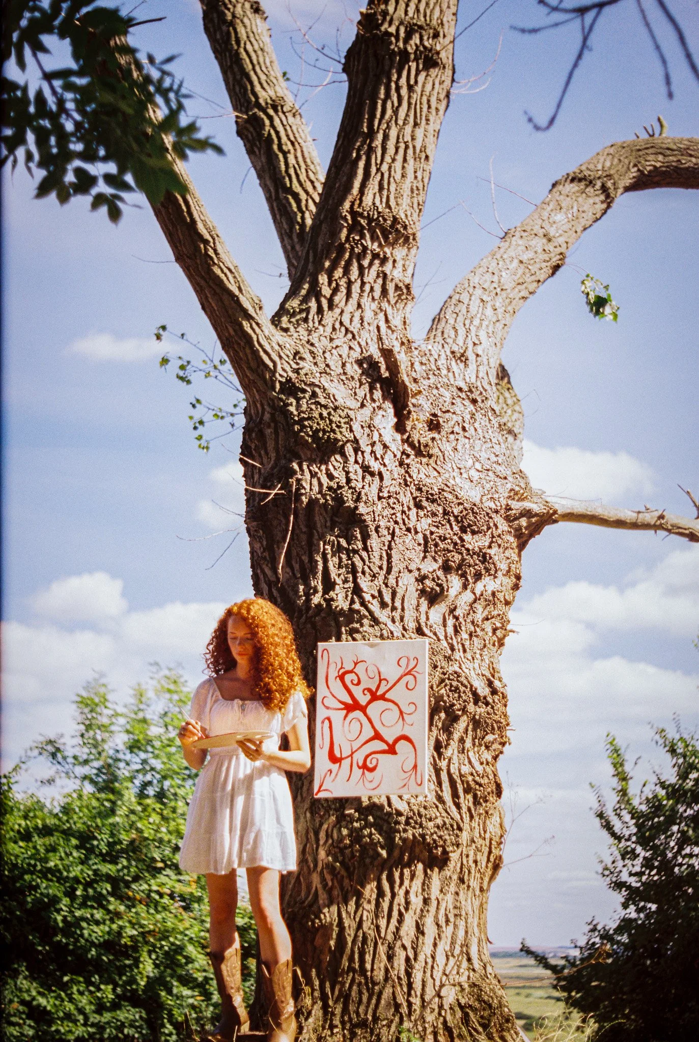 A woman with long curly red hair wearing a white dress and cowboy boots is standing next to a large tree, holding a notebook. There is a white sign with red designs attached to the tree.