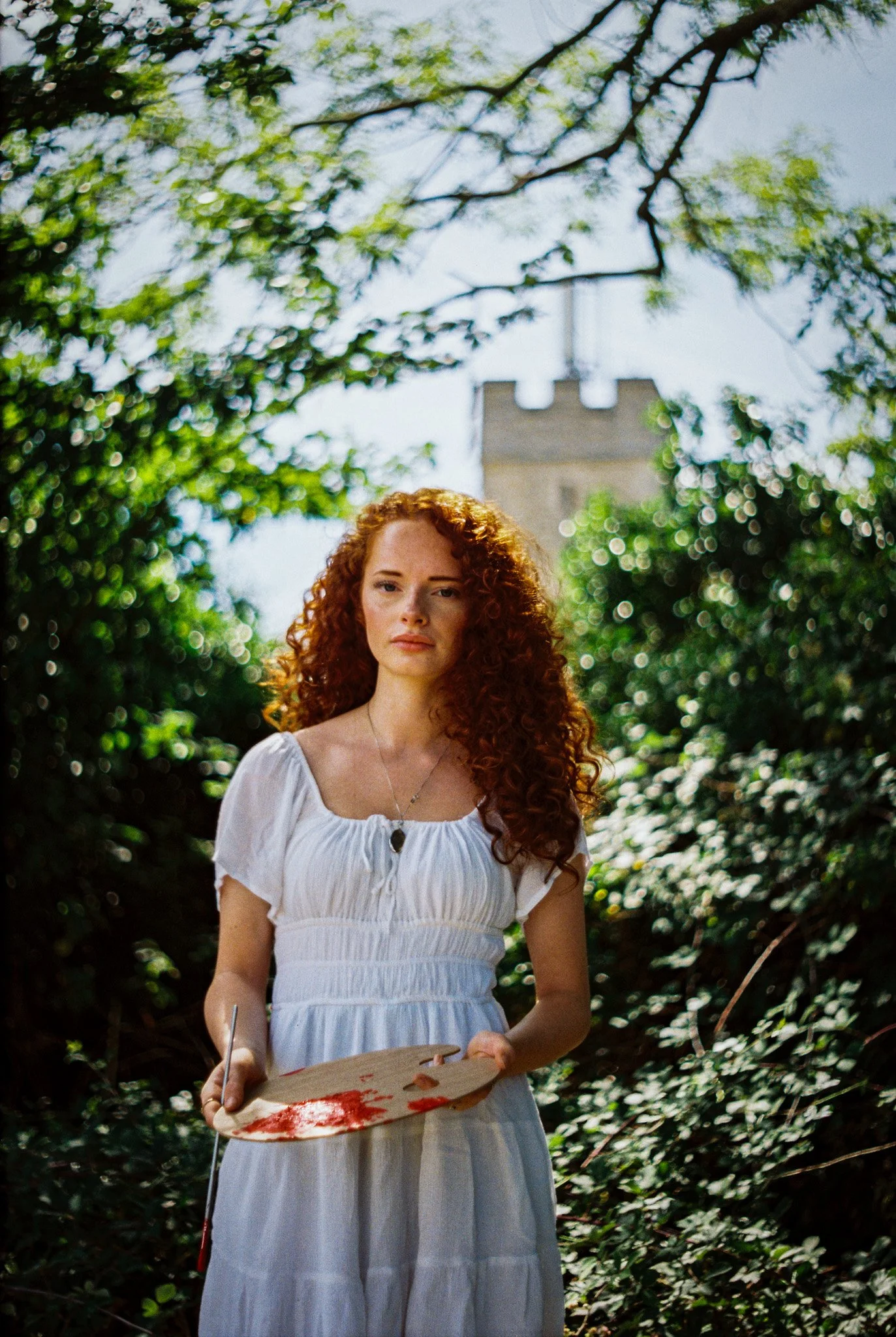A young woman with long curly red hair wearing a white dress standing outdoors among trees, holding a painter's palette with red paint and a paintbrush, with a castle tower visible in the background.