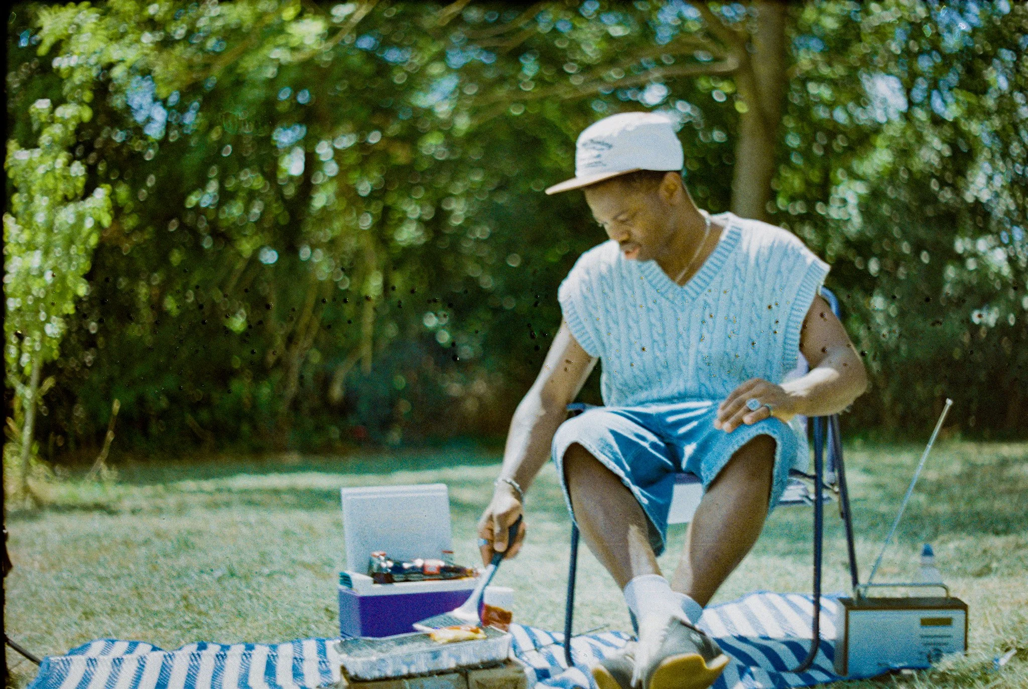 A person sitting outside on a lawn, wearing a large white cap, a sleeveless sweater, and shorts, arranging items on a small table with a striped cover, with greenery in the background.