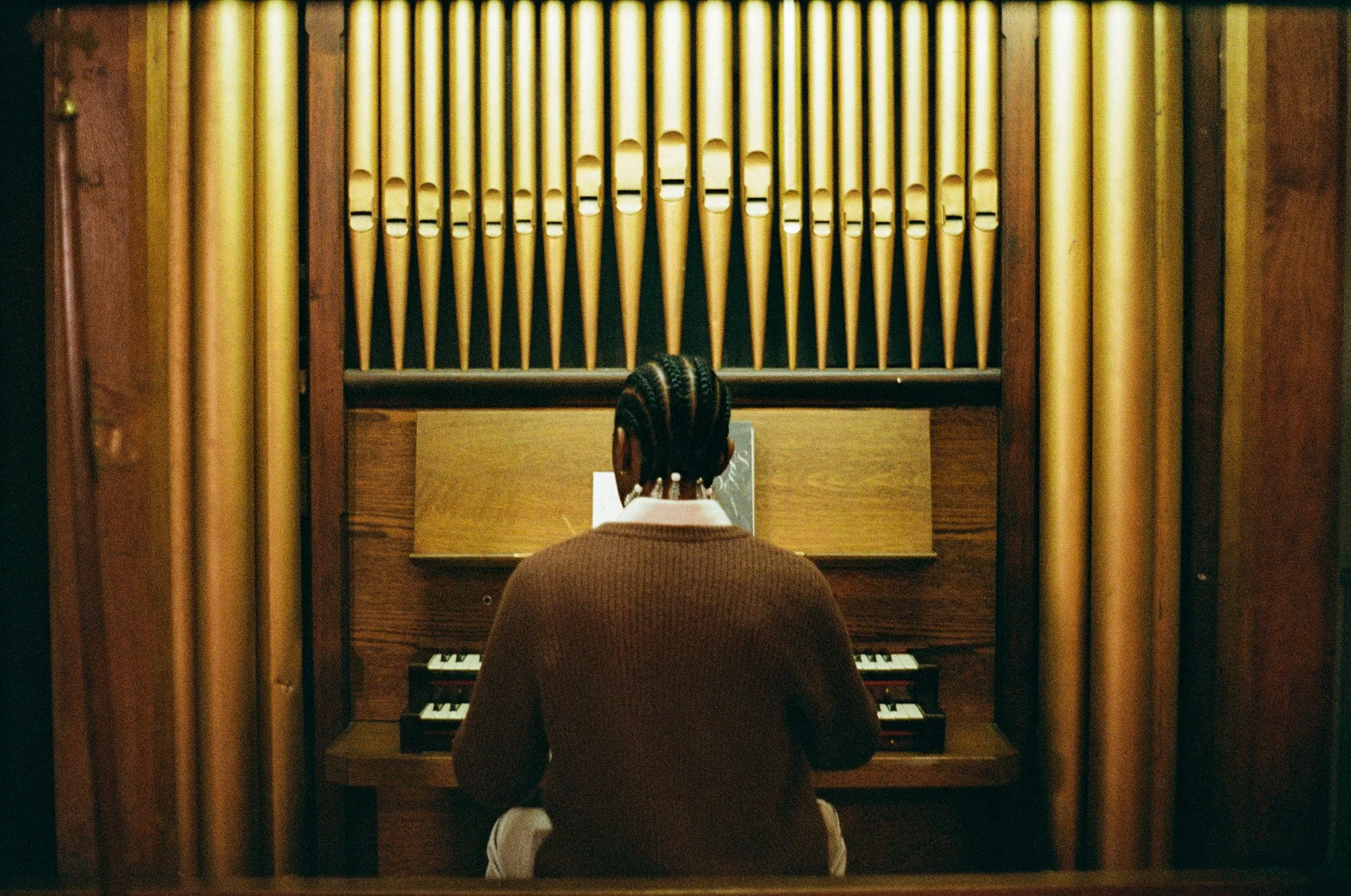 Person with braided hair wearing a brown sweater playing an organ in a music room.