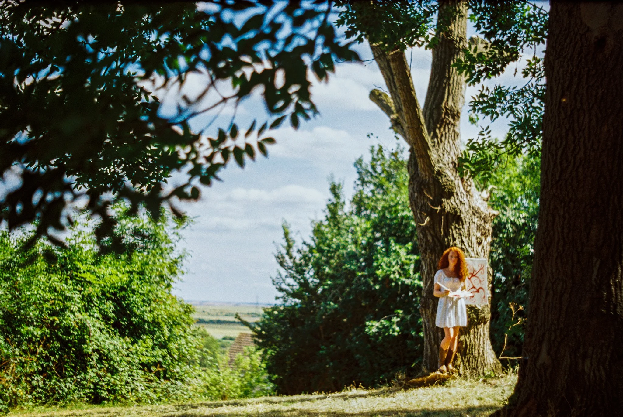 A woman with curly red hair wearing a white dress holding a sign with red writing, standing behind a tree in a lush green outdoor setting.