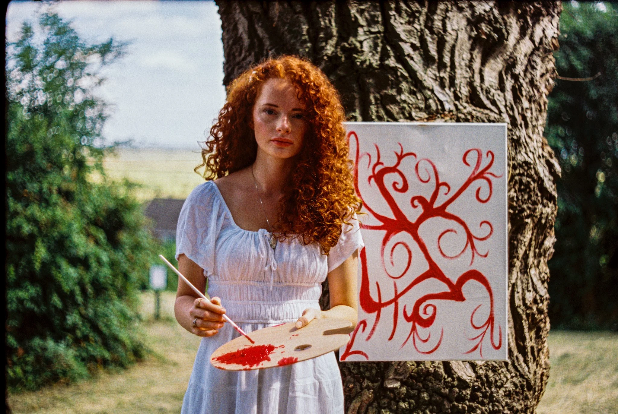 A woman with long curly red hair standing outdoors next to a tree, holding a paint palette and brush, painting a sign with red decorative writing.
