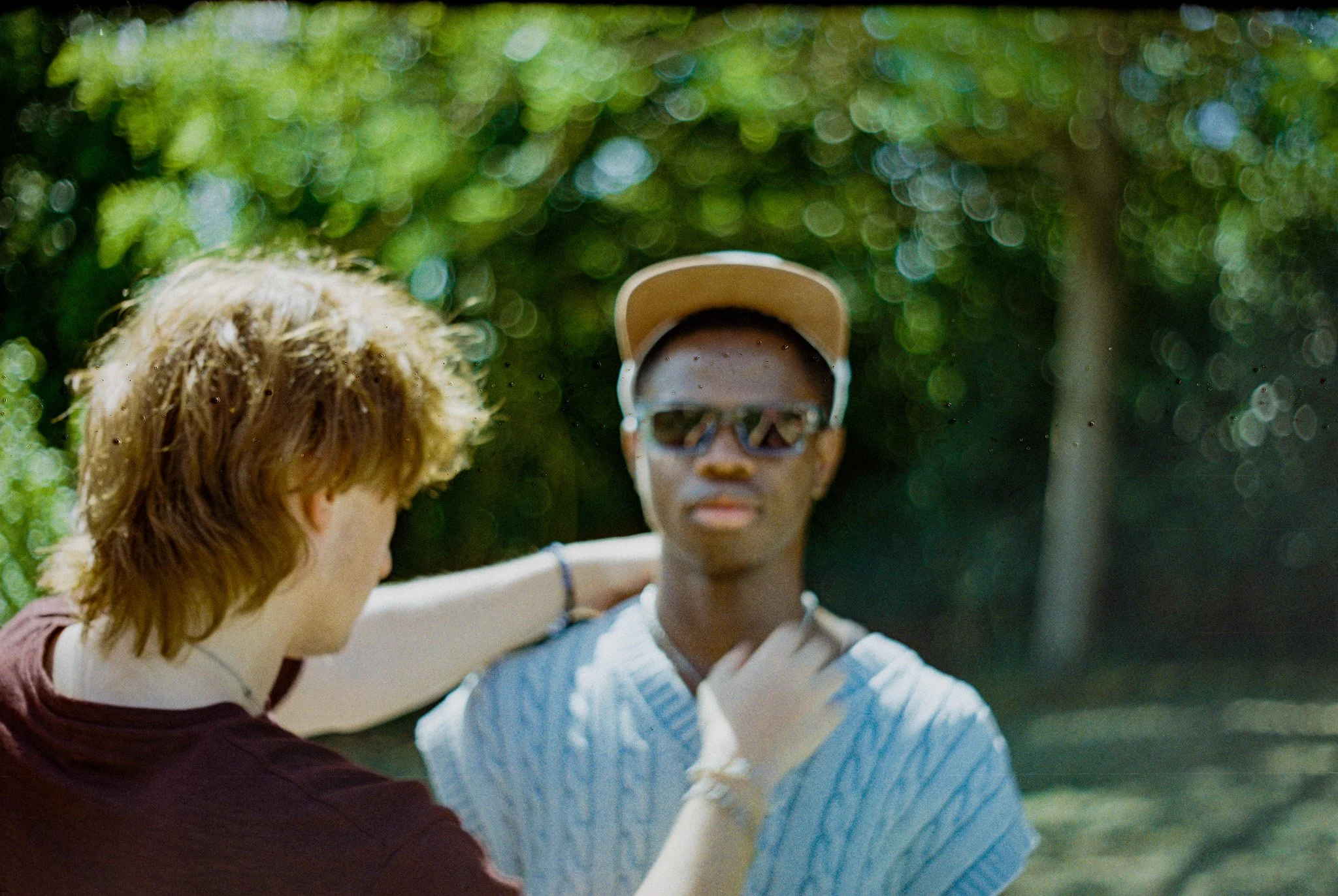 A person with blonde hair and a maroon shirt is hugging a mannequin wearing a beige cap and sunglasses. The person is seen from behind, while the mannequin's face and upper body are visible, with a background of green foliage.