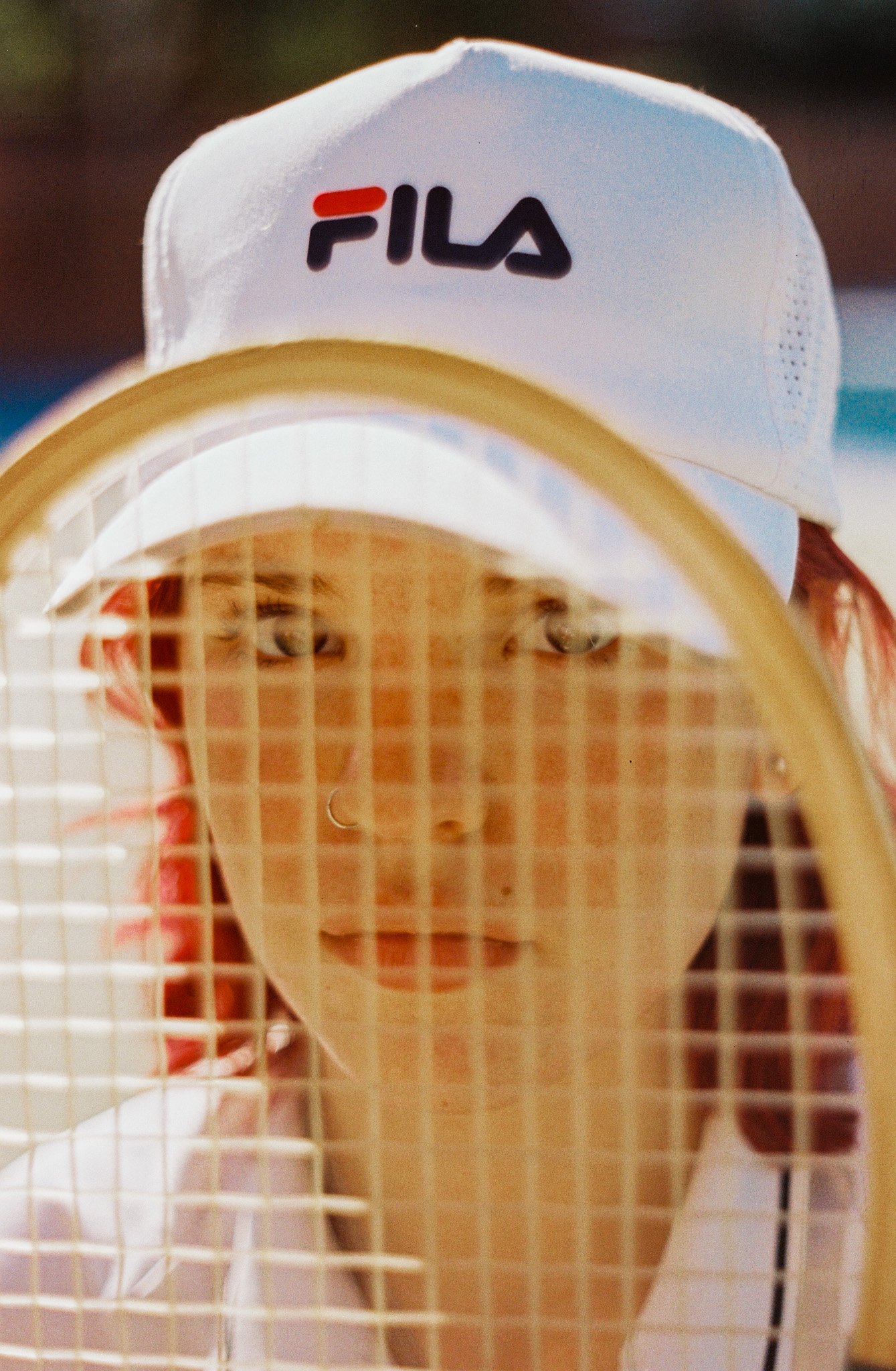 Close-up of a person holding a tennis racket behind a tennis net, with a white FILA baseball cap worn backward.