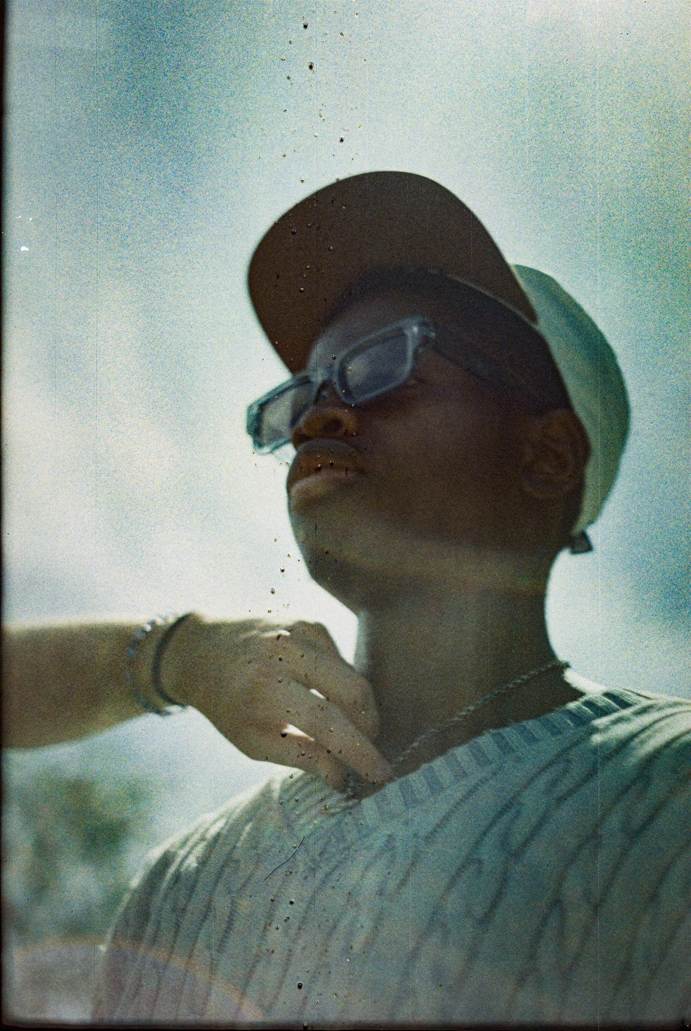 A woman wearing sunglasses, a light-colored cap, a white sweater, and a striped shirt, with her hand resting on a reflective glass surface. The photo is taken from below, showing the sky in the background.