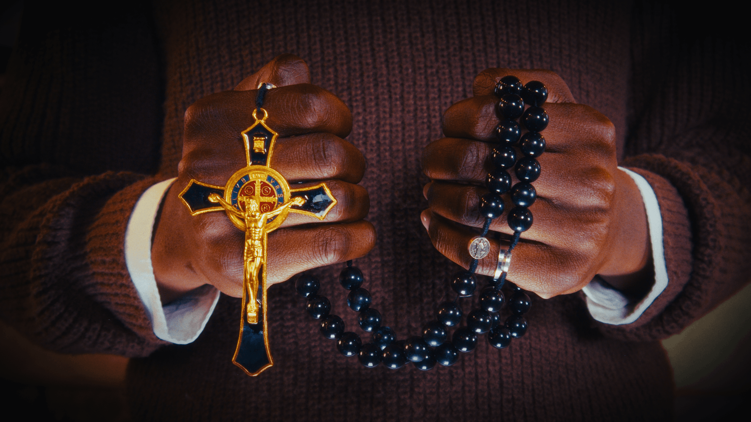 Person holding a gold crucifix and black rosary beads in both hands, wearing a brown sweater.