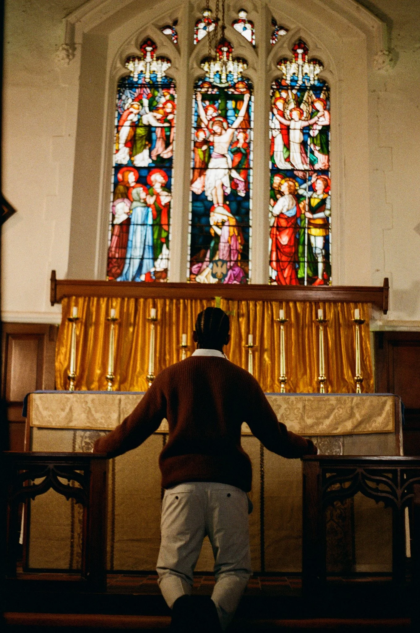 A person standing at an altar in a church, facing a large stained glass window depicting a religious scene with figures, including Jesus on the cross, with a church setting.