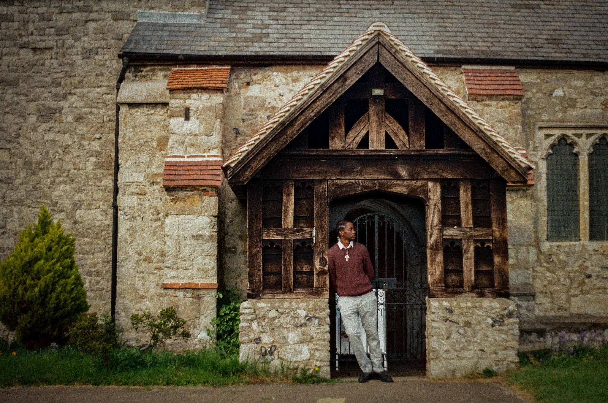 A person standing in front of an old stone building with a wooden entrance frame and a gated door, greenery at the side, and a window in the background.