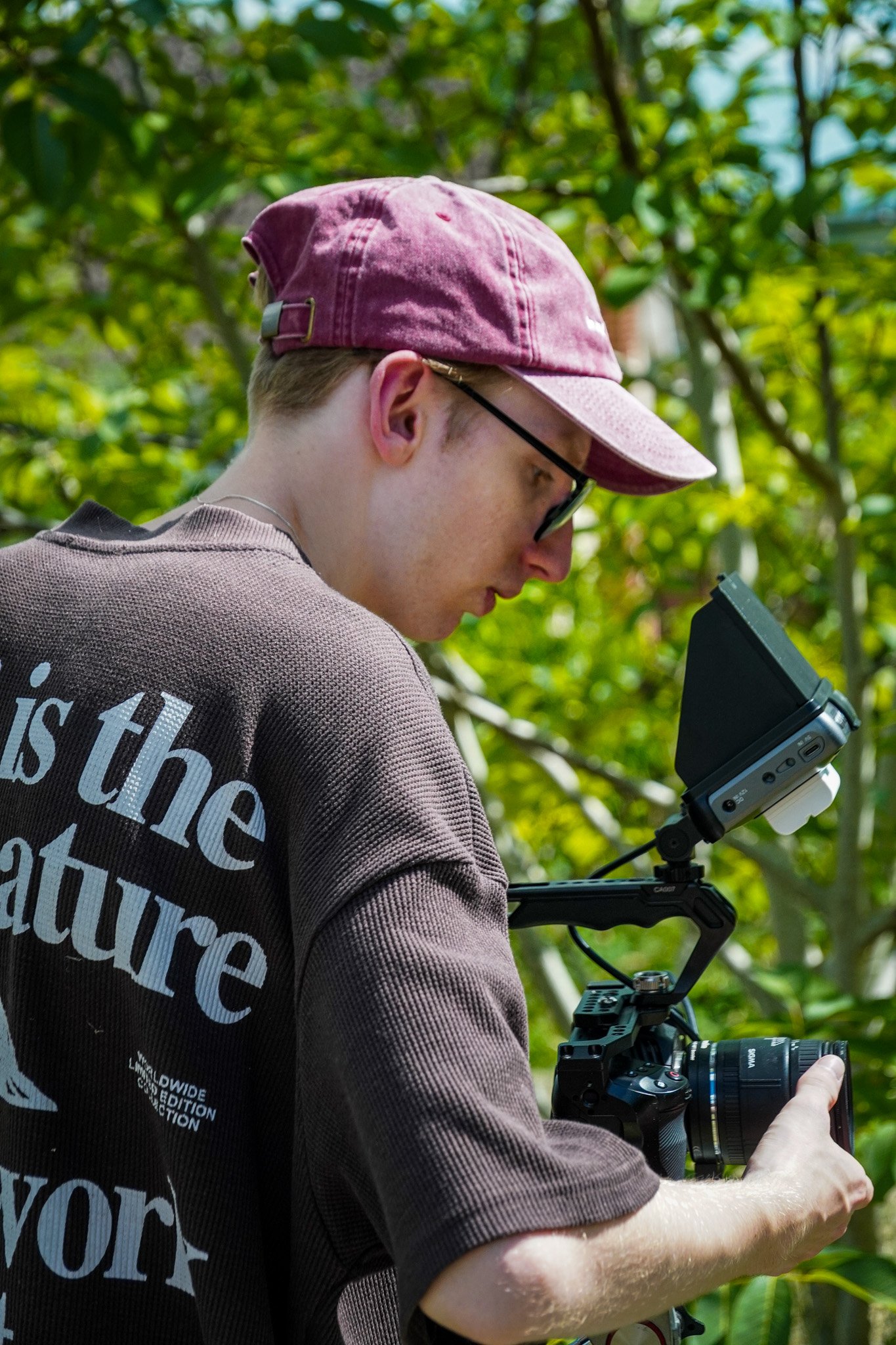 Sam Moore looking at a camera with a viewer screen, outdoors with green foliage in the background.