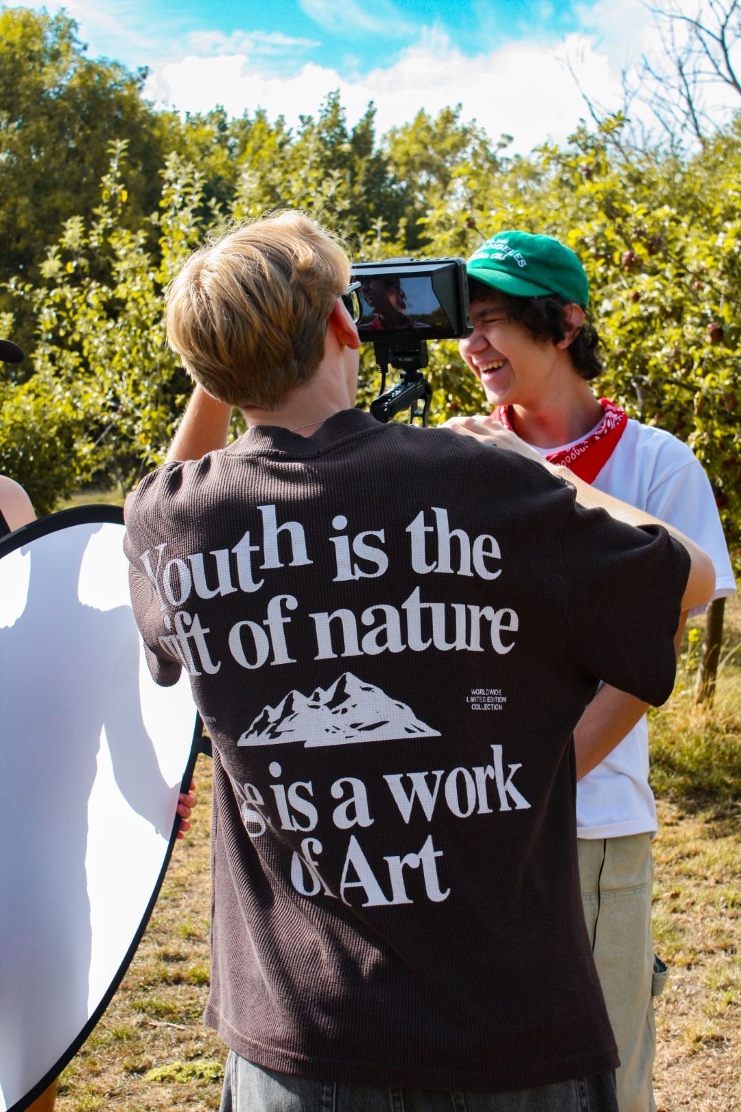 Sam Moore and an actor outdoors, Sam directing and filming the other, who is smiling and wearing a green cap and bandana, with trees and blue sky in background.