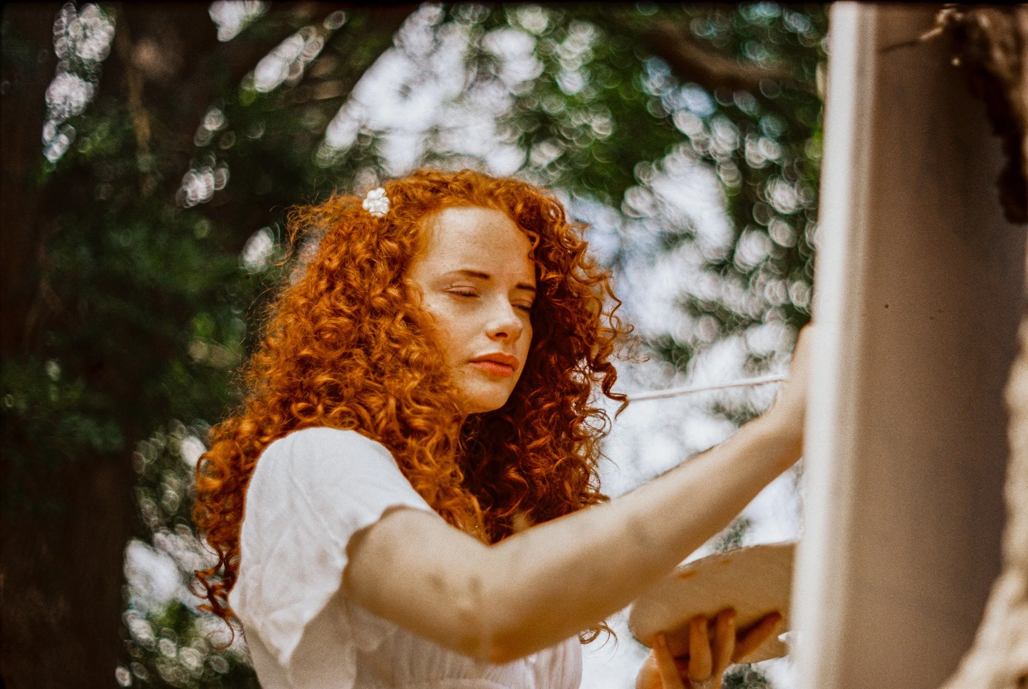 A young woman with curly red hair and freckles is taking a selfie outdoors, with trees and bokeh effects in the background.