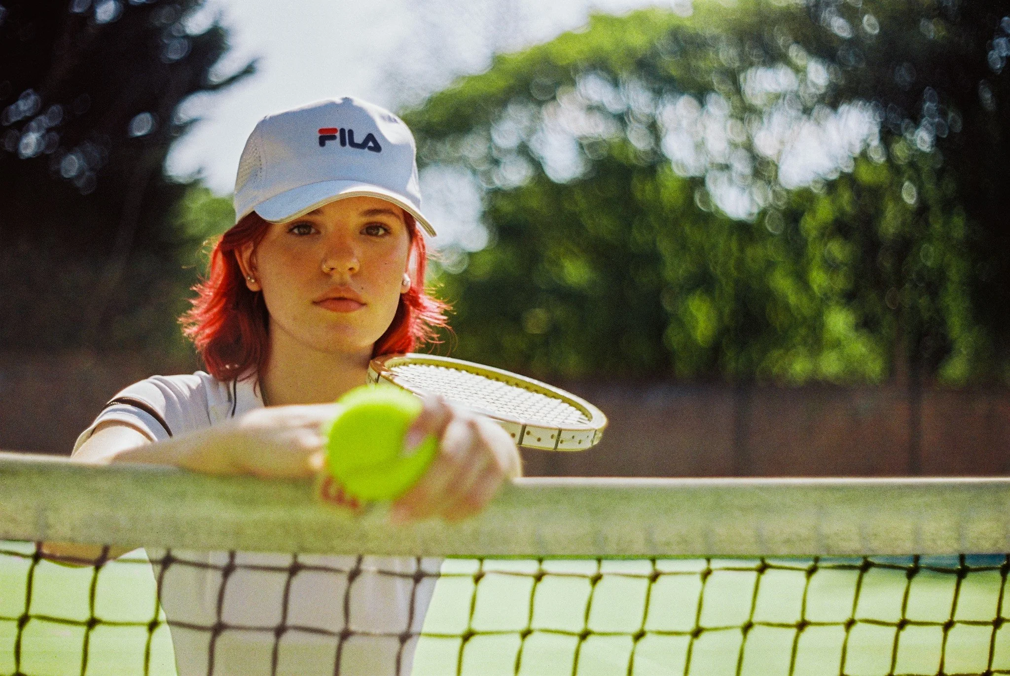 A young woman with red hair and a white hat with the Fila logo is playing tennis on a court, reaching towards the camera with a tennis ball and racquet.